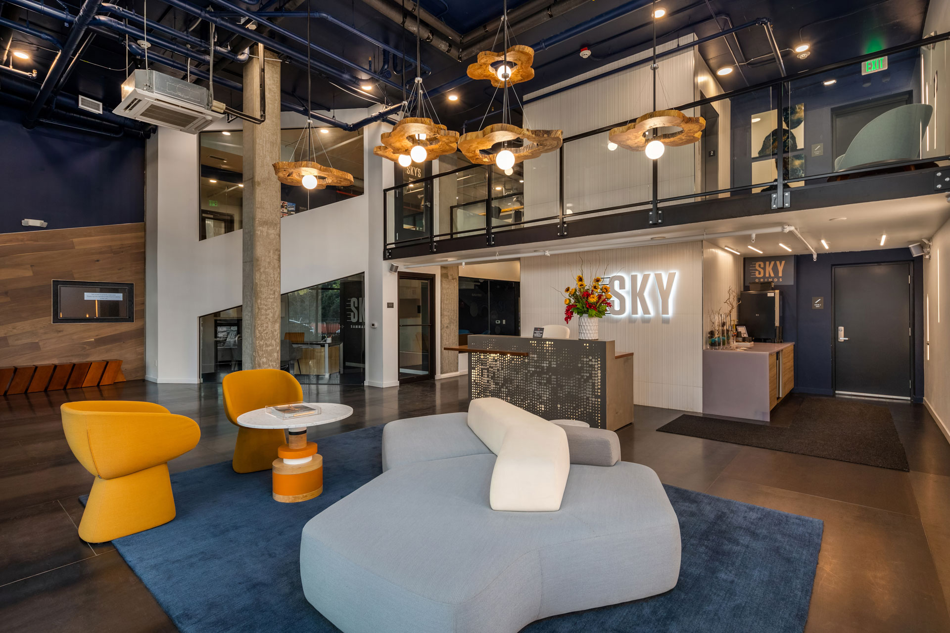 Modern office lobby with contemporary furniture including an inviting gray sofa, yellow armchairs, and a circular coffee table. Reception desk with a lit "SKY" sign in the background. Mezzanine level above, stylish pendant lighting, and minimalistic decor throughout.