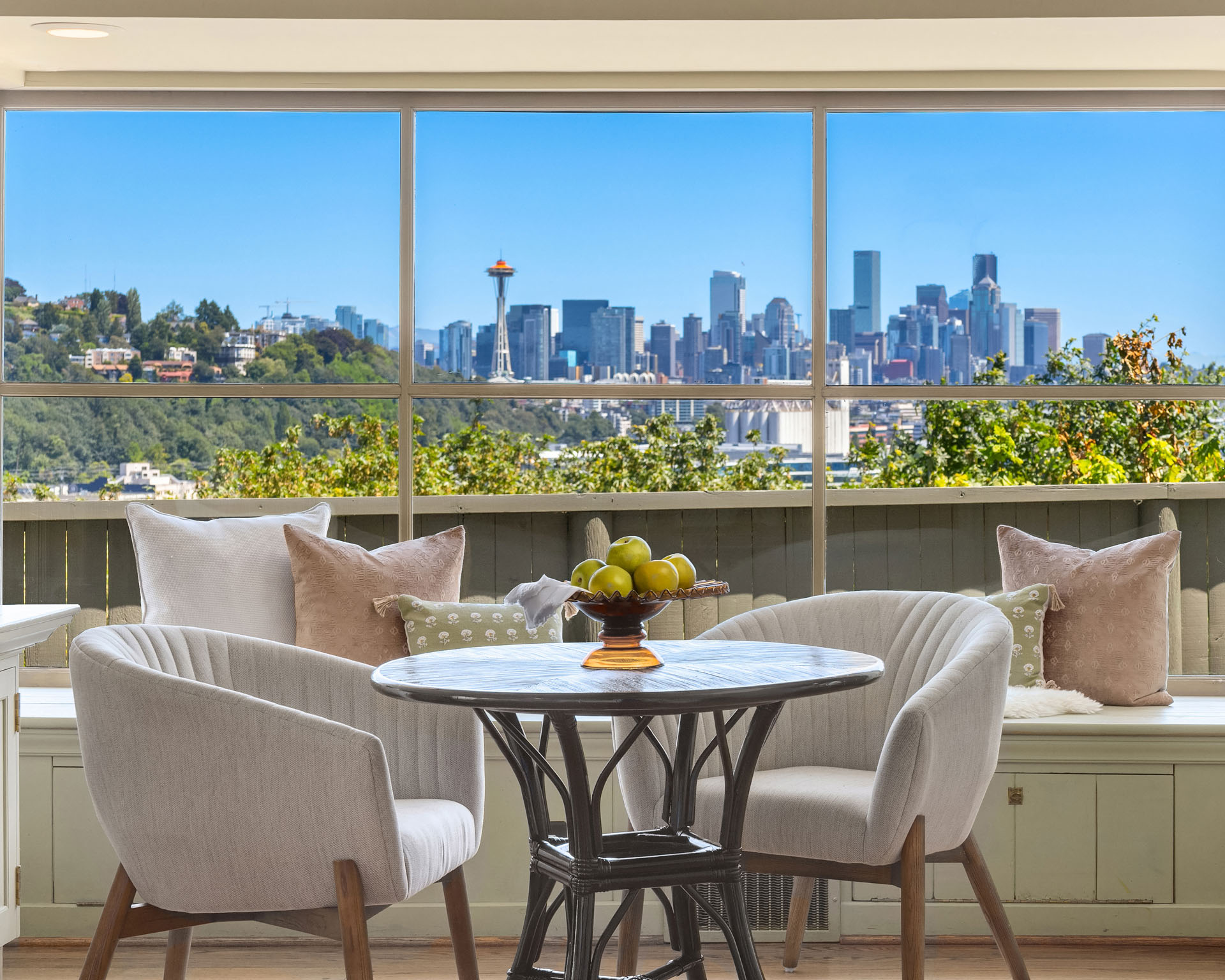 A cozy patio area with a round table and two cushioned chairs overlooks a stunning cityscape. The table holds a bowl of green apples. Large windows and greenery enhance the setting, while a clear blue sky and notable skyline, including a prominent tower, are visible.