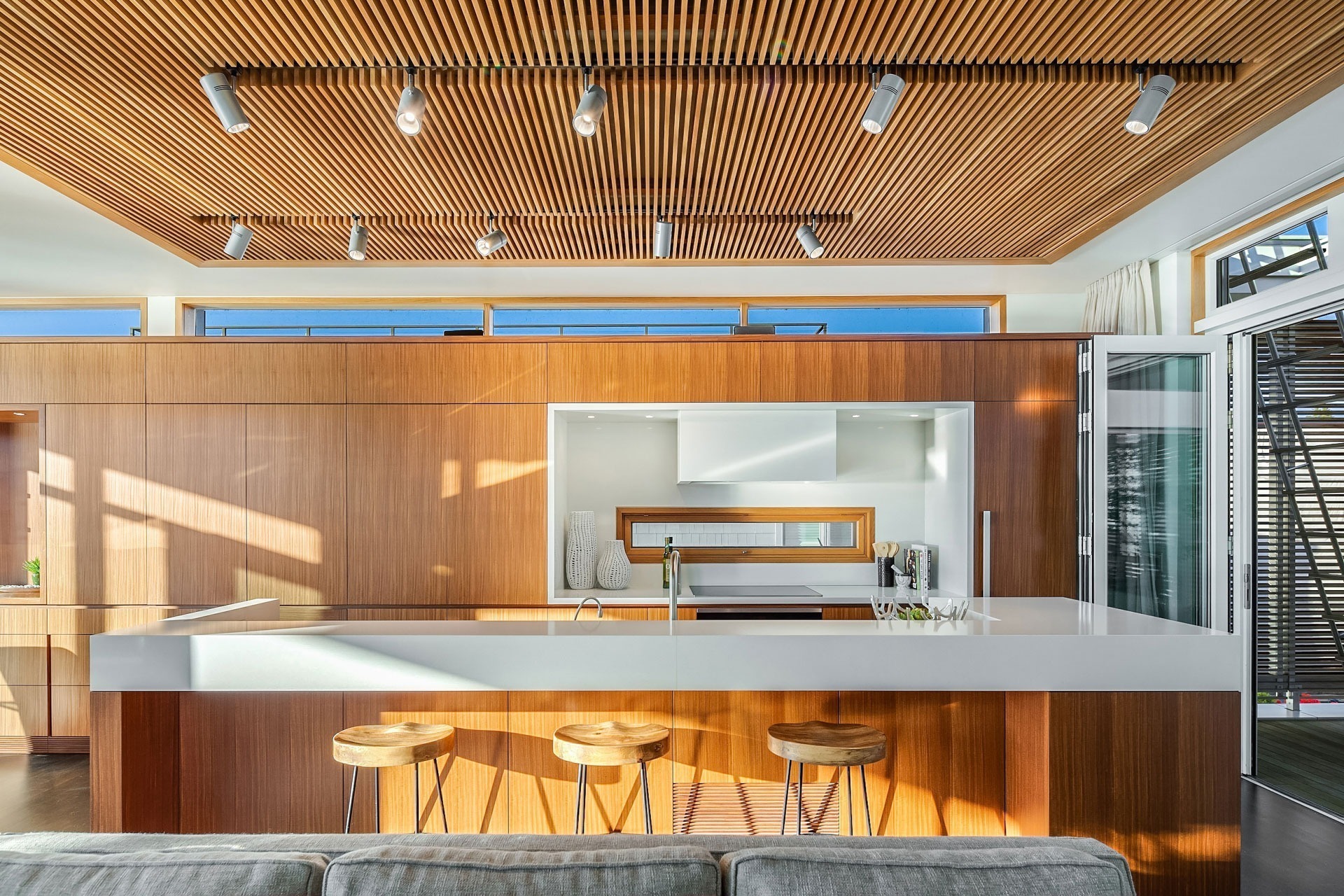 A modern kitchen featuring a sleek, white island with three wooden bar stools underneath. The island faces a wooden wall with built-in appliances and a white countertop. The ceiling is adorned with wooden slats and track lighting. Large windows bring in natural light.