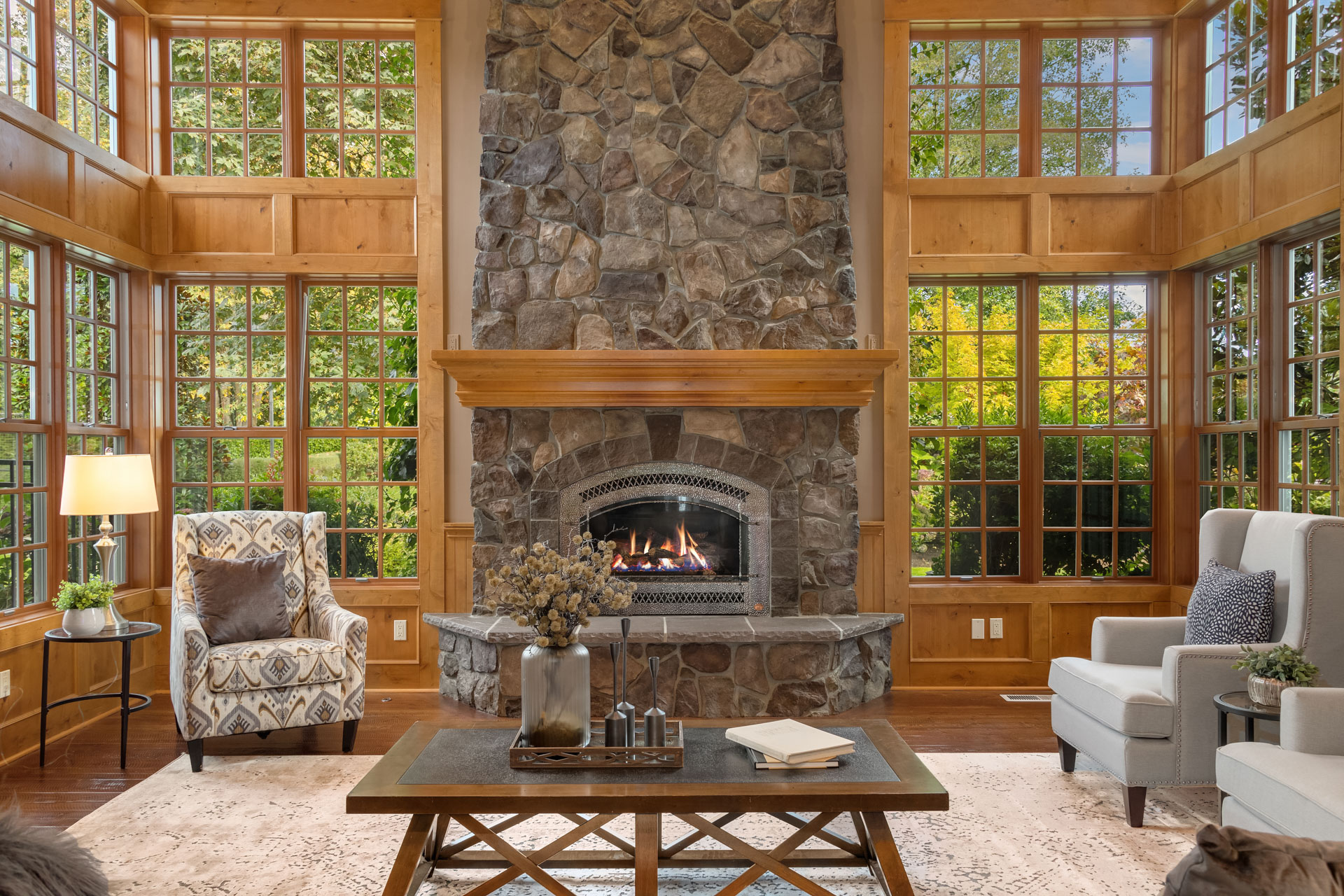 A cozy living room features a large stone fireplace with a wooden mantel. Tall windows surround the room, letting in ample natural light. The space is furnished with patterned and plain armchairs, a central coffee table, and a floor lamp. Nature is visible outside.