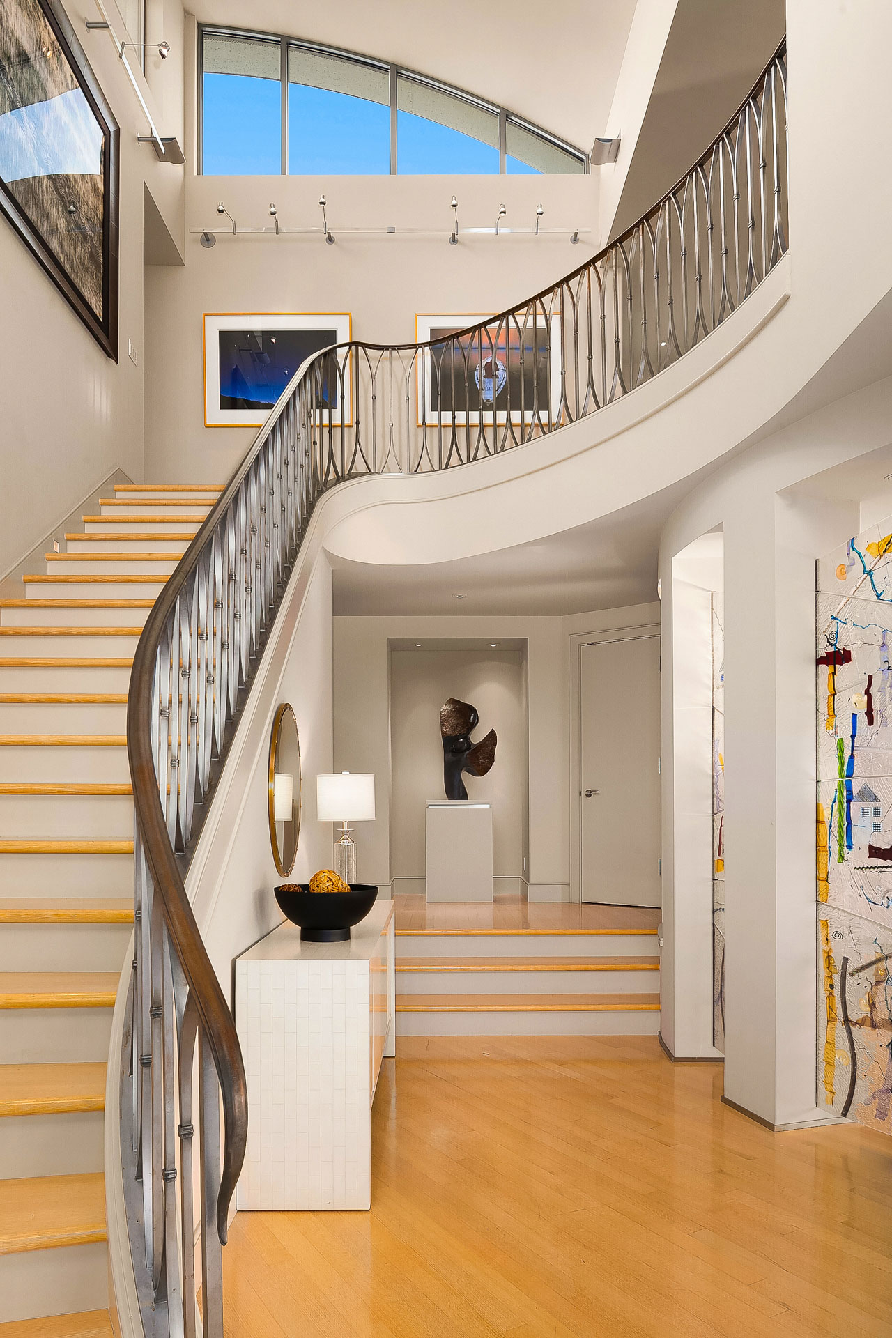 A modern, well-lit foyer with a curved staircase featuring a sleek metal railing. The space has light wooden floors, abstract art on the walls, and a sculpture on a pedestal near the entrance. A round mirror and table lamp adorn a console table at the bottom of the stairs.