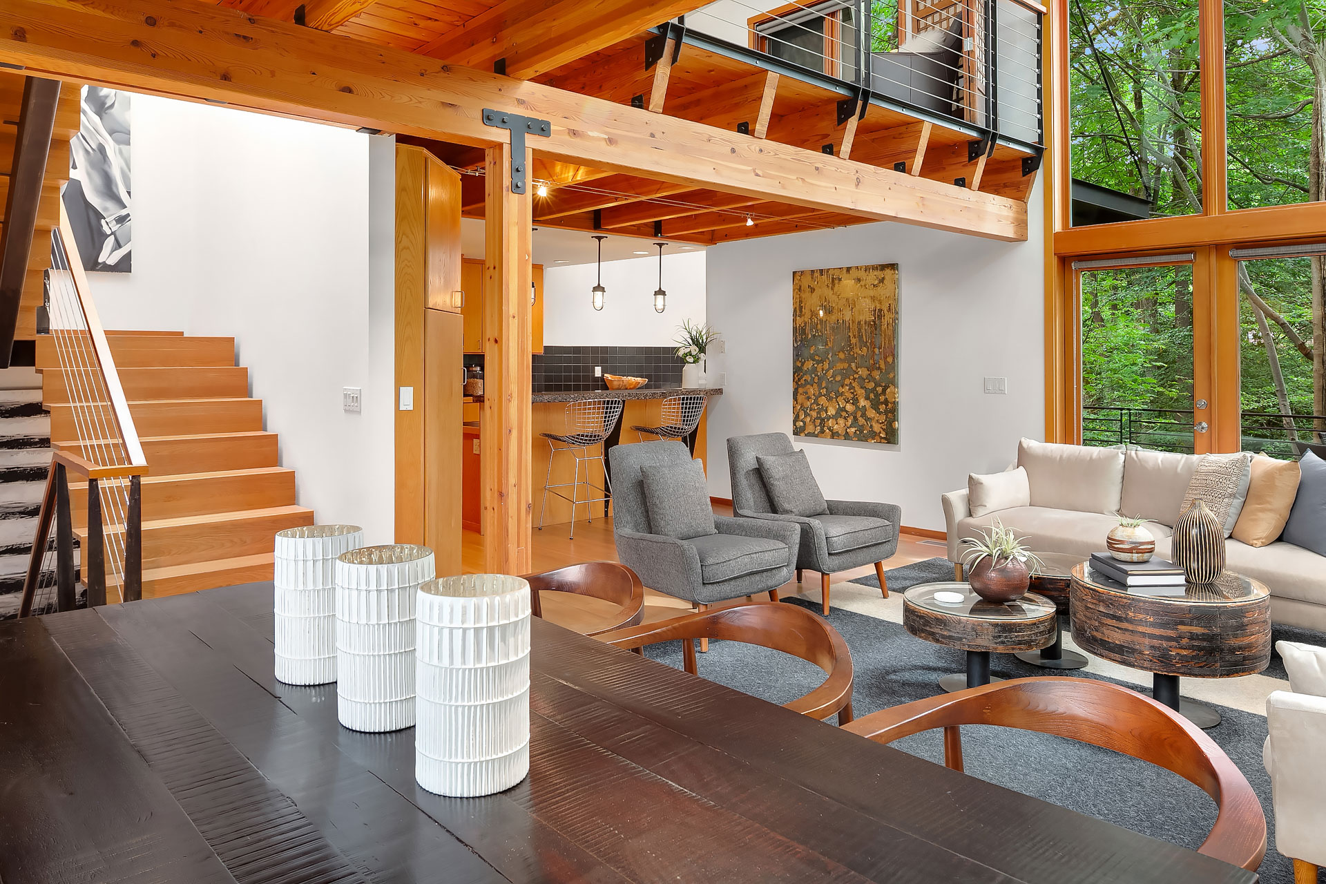 Modern living room with an open concept design featuring wooden beams, a staircase with wooden steps, and a mix of modern furniture including gray armchairs, a dark dining table, and wooden chairs. The room has large windows overlooking greenery, and a contemporary kitchen in the background.