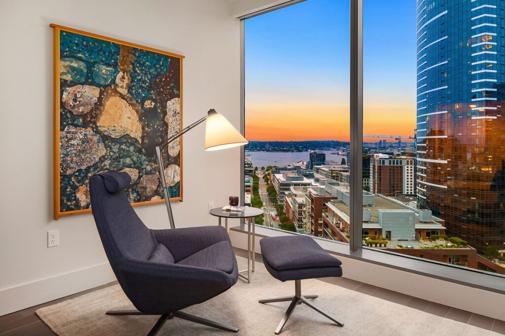 A modern lounge area features a dark blue chair with a matching ottoman, positioned next to a sleek floor lamp and a small side table. A large abstract painting hangs on the wall. A large window showcases a cityscape at sunset, with buildings and a body of water visible.