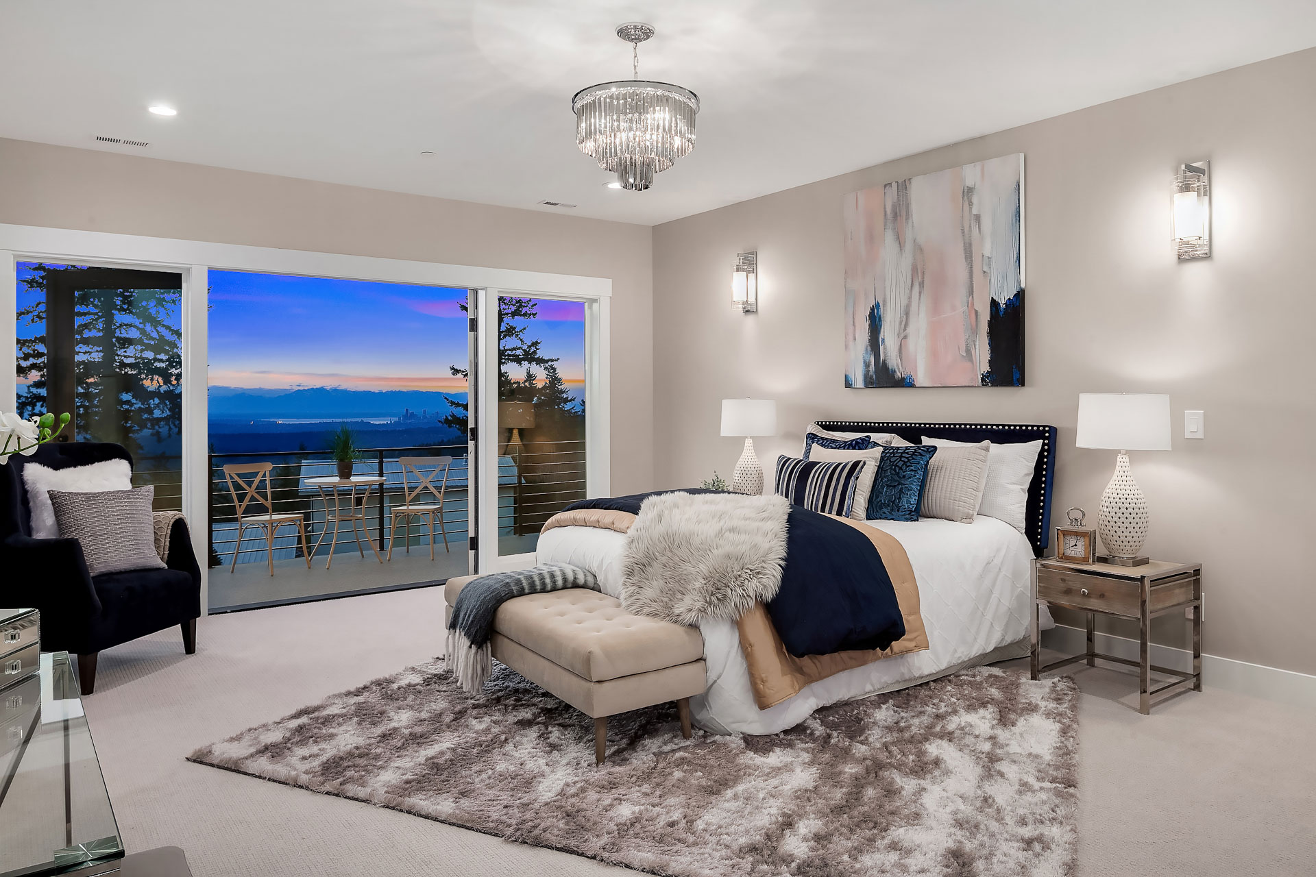 Modern bedroom with a large bed adorned with plush pillows and a faux fur throw. A bench sits at the foot of the bed. The room features a chandelier, an abstract painting, and a shaggy rug. Sliding glass doors open to a deck with a view of trees and mountains at sunset.