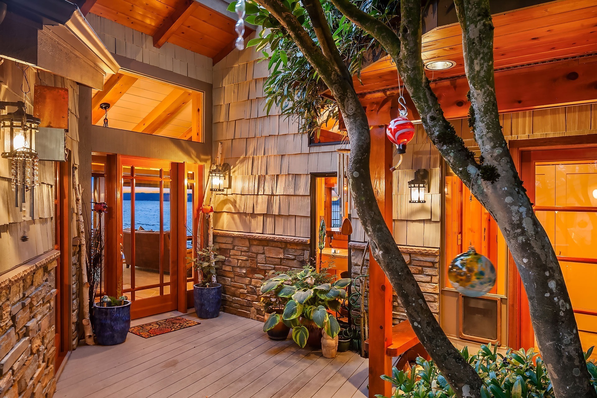 A warmly-lit, rustic wooden porch at dusk features a gabled roof, shingle siding, and stone accents. Potted plants and hanging ornaments decorate the space. Large windows and a glass door offer a glimpse into the cozy interior, with a peaceful lake visible in the background.