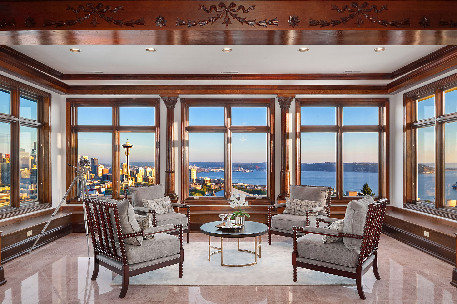 A luxurious living room with large windows offering a panoramic view of a cityscape and waterfront. The room features elegant wooden trim, beige armchairs with gray cushions, a round coffee table with a flower arrangement, and decorative ceiling moldings.