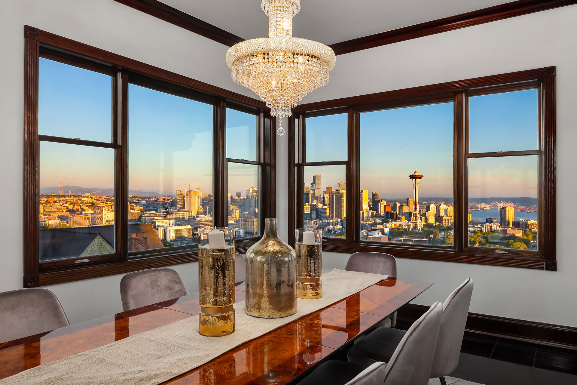 A dining room with a wooden table, gray chairs, and an elegant chandelier. The large windows provide a panoramic view of a city skyline during sunset, featuring a famous observation tower and various high-rise buildings. Decorative vases are on the table.