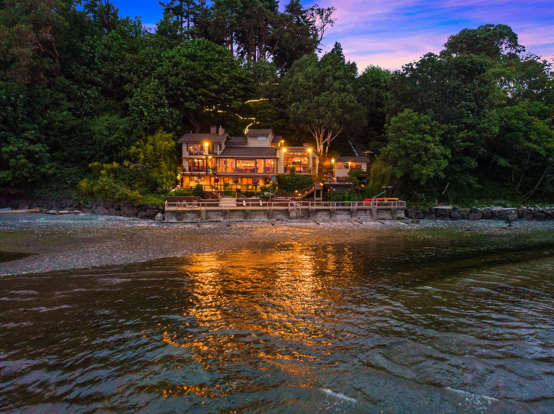 A cozy, well-lit house is nestled among dense trees on a shoreline at dusk. The glow from the house reflects off the calm water, creating a warm and inviting ambiance. The sky is a gradient from blue to purple, enhancing the tranquil setting.