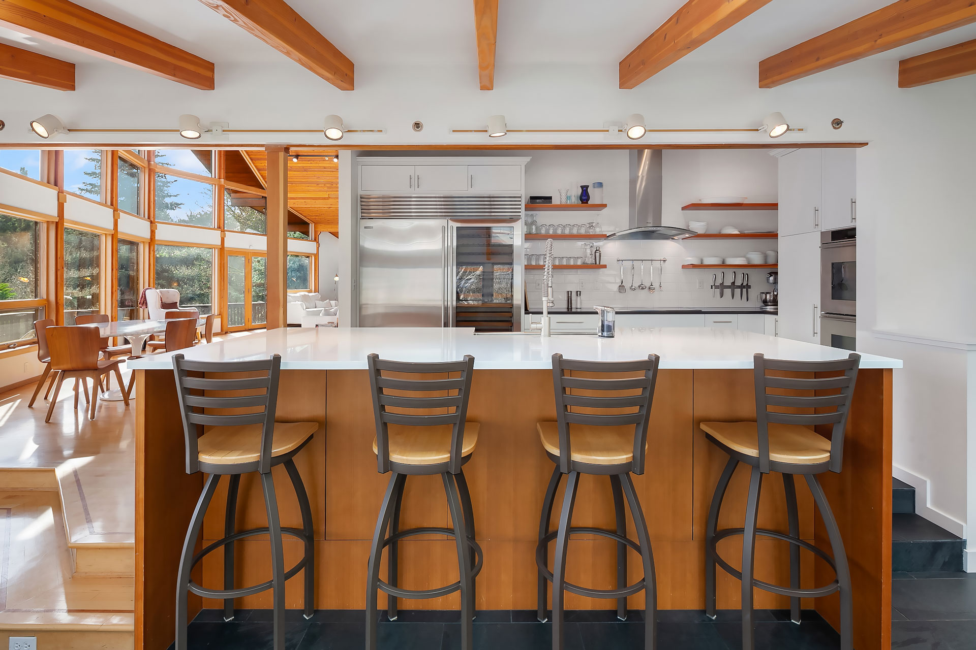 Modern kitchen with a large white island countertop and four wooden bar stools. The space features stainless steel appliances, open shelves, and cabinets. Wooden ceiling beams and large windows bring in natural light, creating an airy and inviting atmosphere.