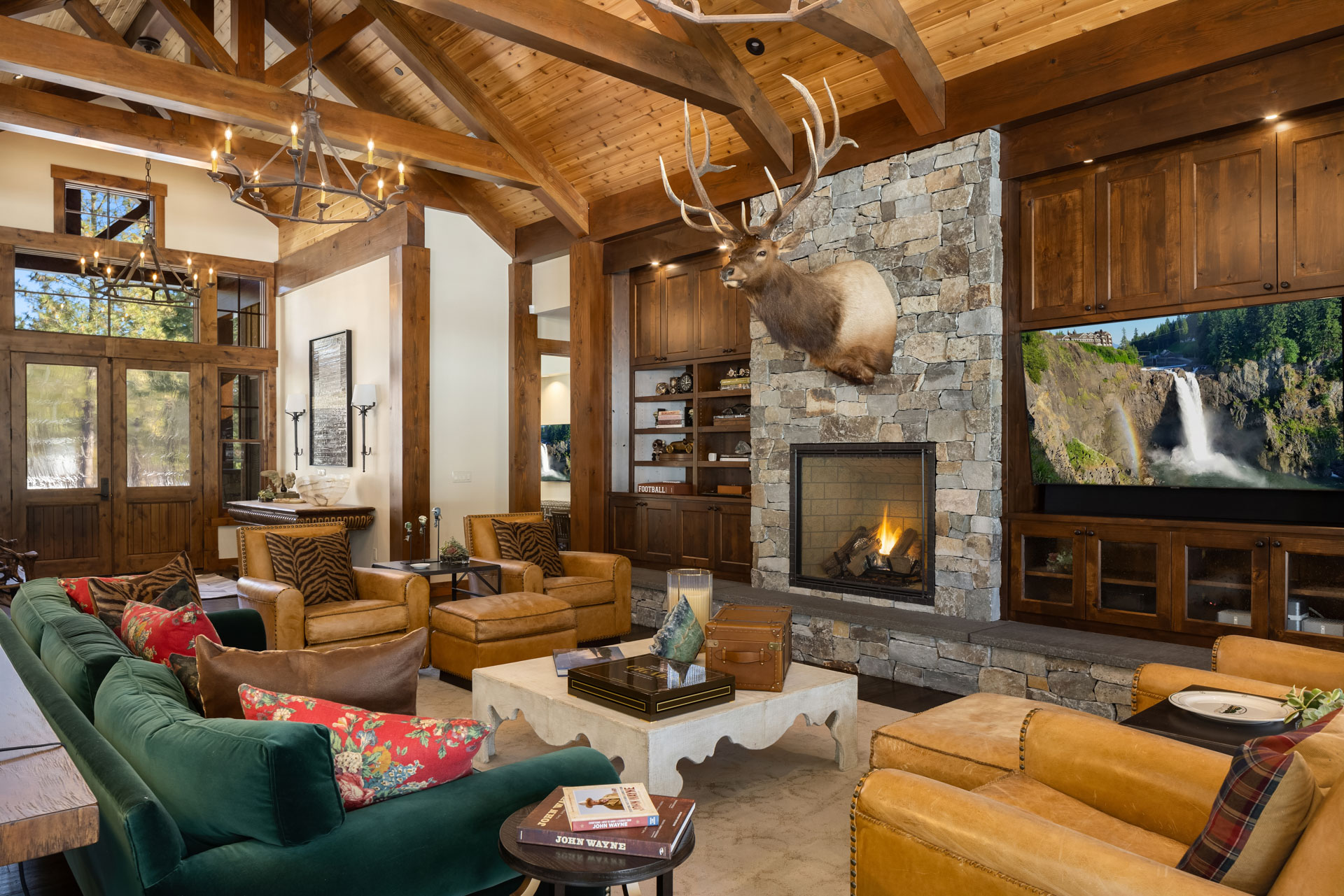 A cozy living room with a rustic wooden beam ceiling features a fireplace with a stone surround and a mounted deer head. Furnishings include green and brown sofas, armchairs, and a coffee table. Large windows offer a view of a waterfall and lush greenery outside.