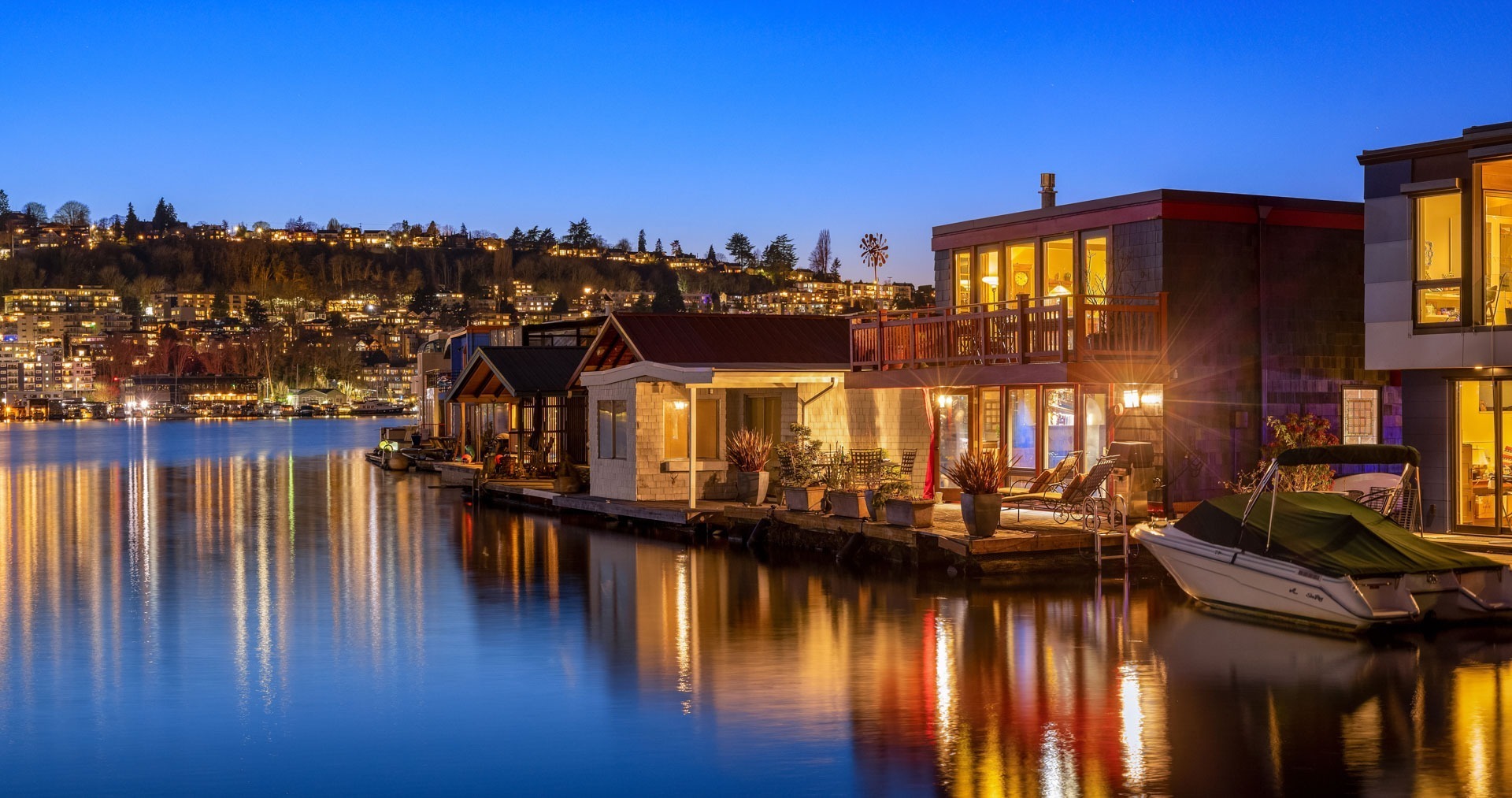 A serene evening view of houseboats reflecting on a calm lake under a clear, deep blue sky. Warm lights from the houseboats and surrounding buildings illuminate the waterfront, casting golden reflections on the water. A small motorboat is moored to a dock on the right.