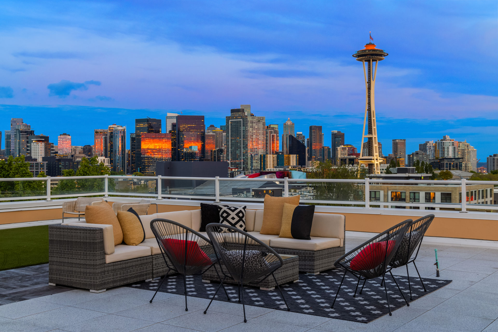 A modern rooftop patio with a stylish seating arrangement overlooks the city skyline at dusk. The scene features cozy chairs, a sectional sofa, and a coffee table on a patterned rug. The iconic Space Needle is illuminated in the background against a colorful sky.