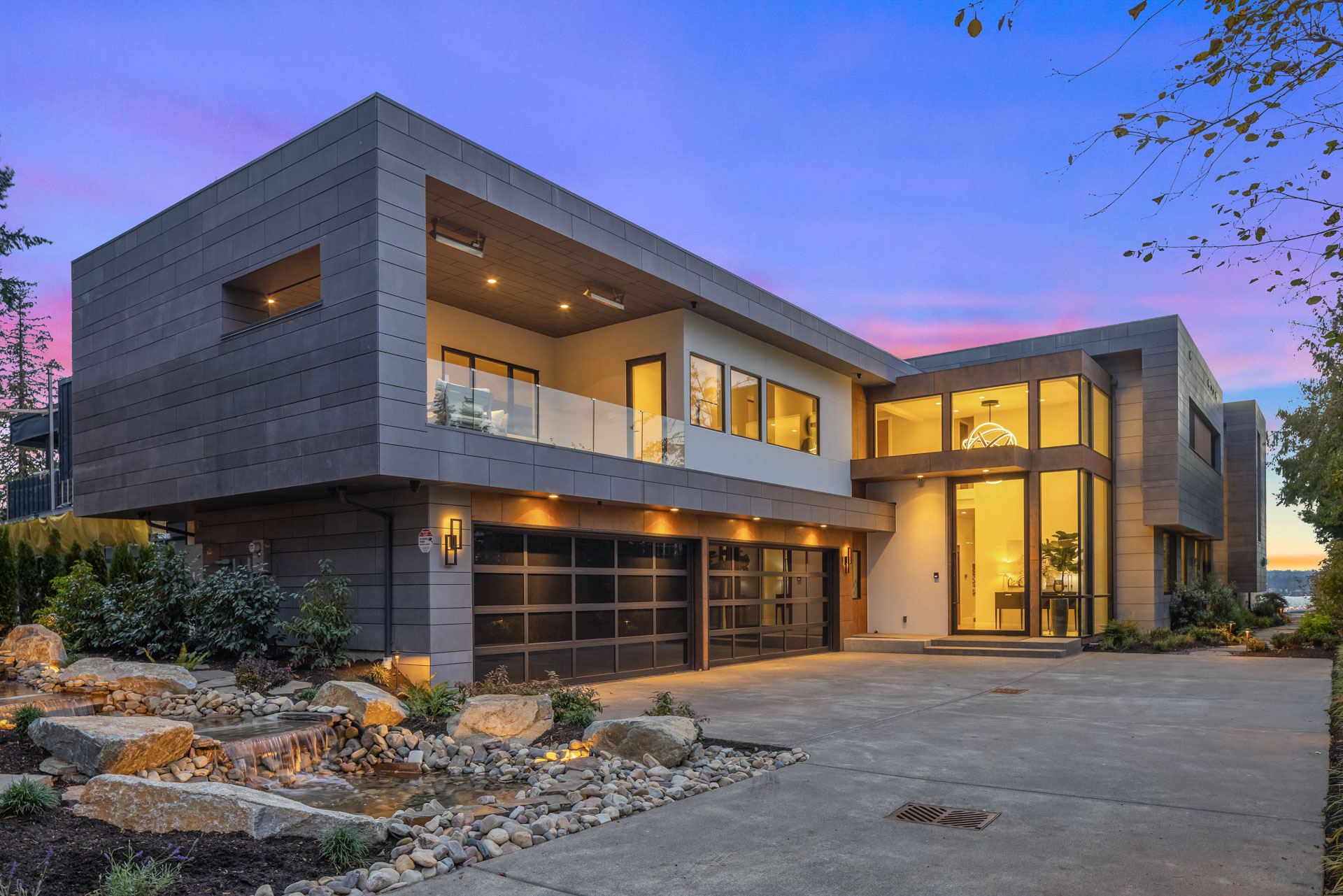 Modern two-story house with a flat roof, large glass windows, and a spacious driveway at sunset. The exterior includes a waterfall feature with surrounding rocks and plants. The home has warm, ambient lighting and a second-floor balcony.