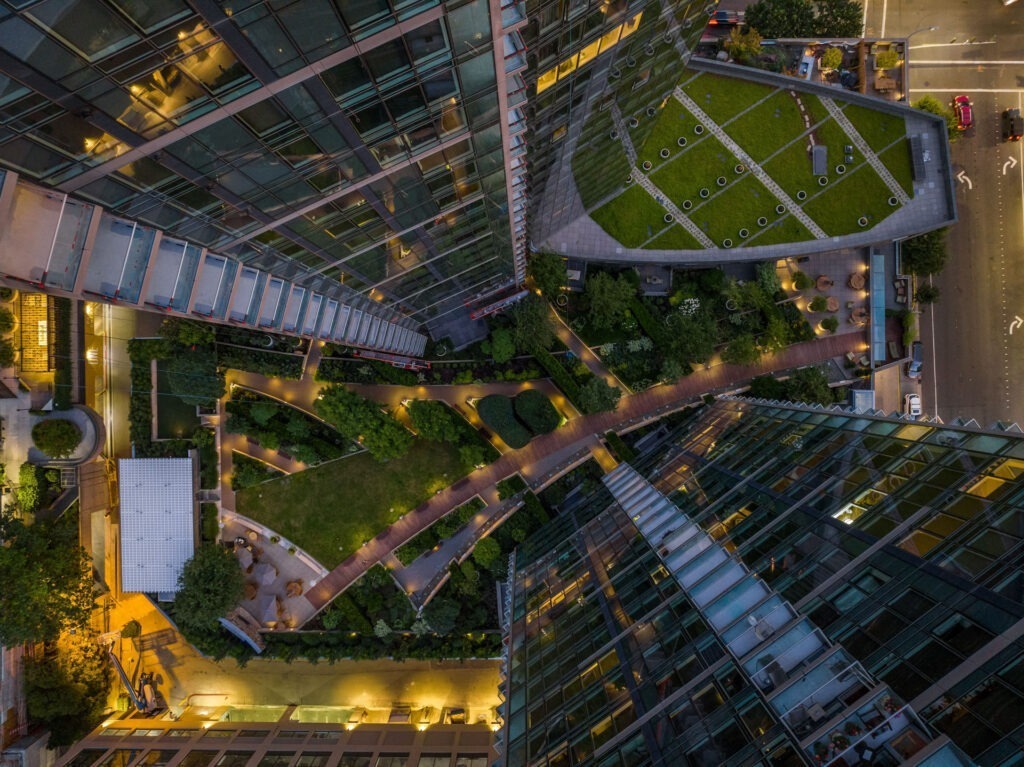 Aerial view of an urban landscape featuring tall glass buildings surrounding a geometric courtyard with landscaped greenery, pathways, and benches. The streets visible at the top right corner are illuminated by streetlights and traffic signals with some cars driving by.