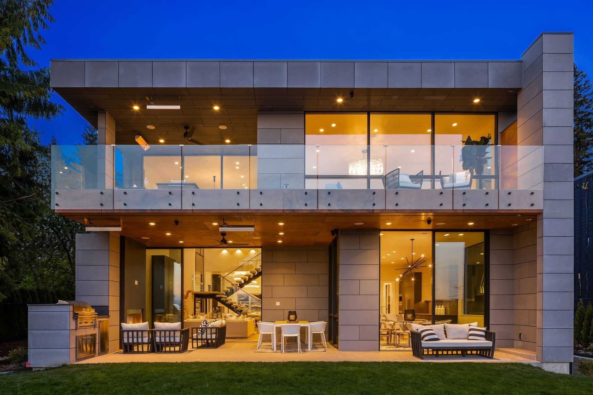 Modern two-story home at dusk featuring large glass windows and a spacious outdoor patio. The ground floor patio has comfortable seating and a grill, while the upper floor has a balcony with glass railings. The interior is lit warmly, highlighting a contemporary design.