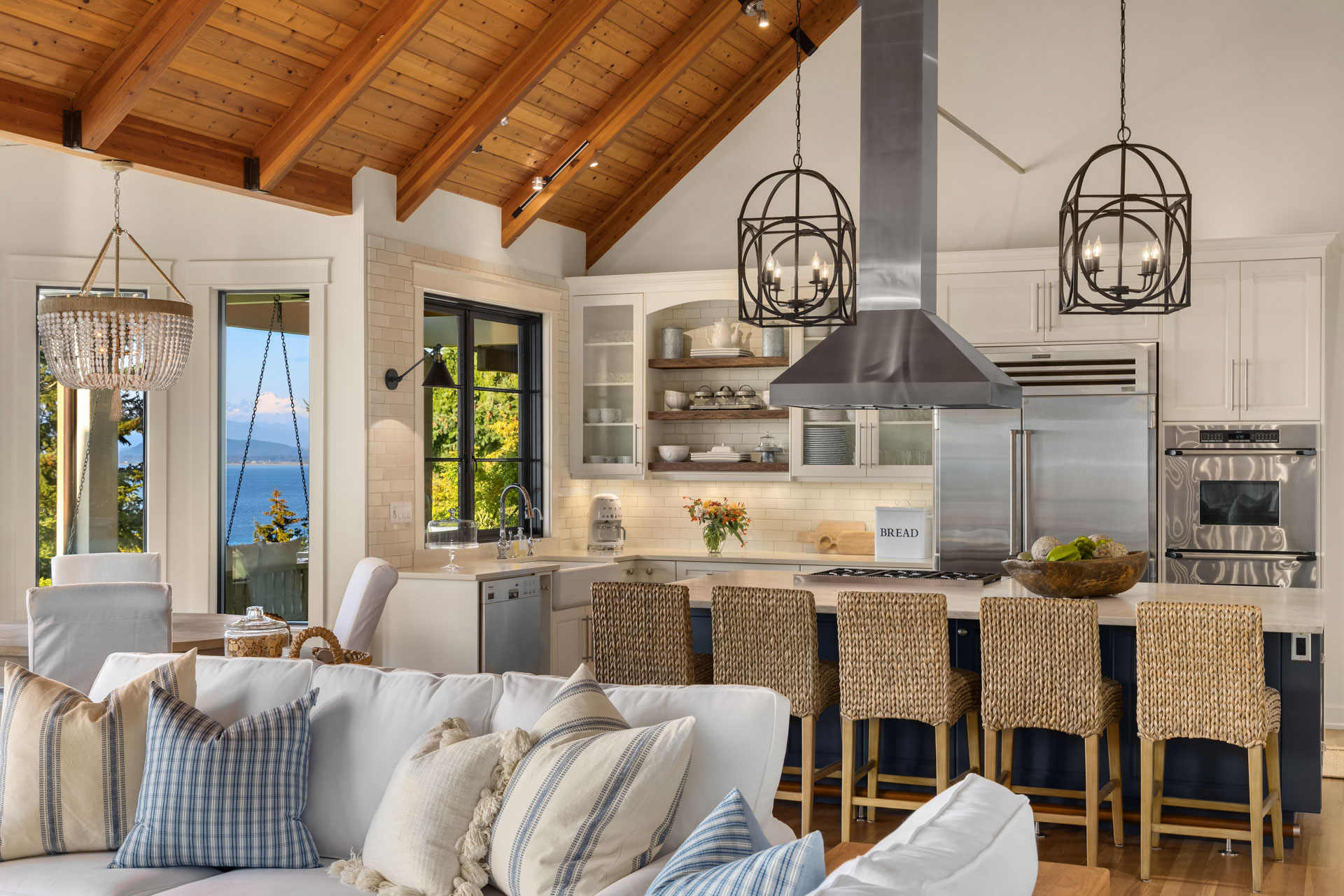 A modern, open-concept kitchen with a high, wooden-beam ceiling. The space includes a large island with wicker barstools, stainless steel appliances, and white cabinets. Large windows offer a scenic view of a lake and mountains, with natural light filling the room.