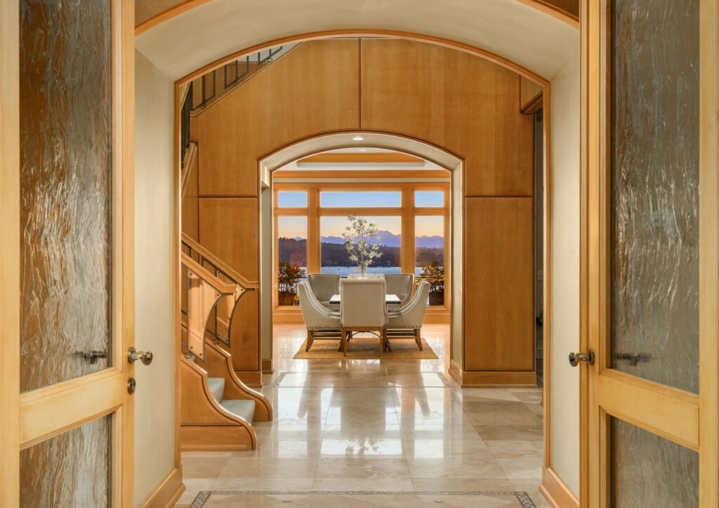 A warm, elegant hallway leads to a cozy living area with a large window offering a stunning sunset view. Wooden walls, a graceful staircase to the left, and a set table with chairs are visible, creating an inviting and luxurious atmosphere.