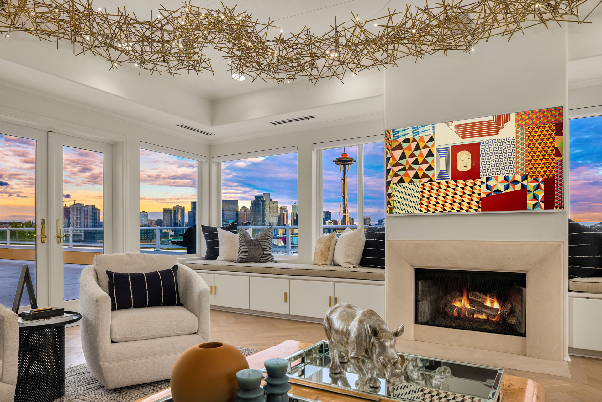 A modern living room with large windows offering a cityscape view, including the Space Needle. The room features a lit fireplace, abstract art above it, cozy armchairs, and modern accents, such as a gold-tone decorative light fixture and sculptures on the coffee table.