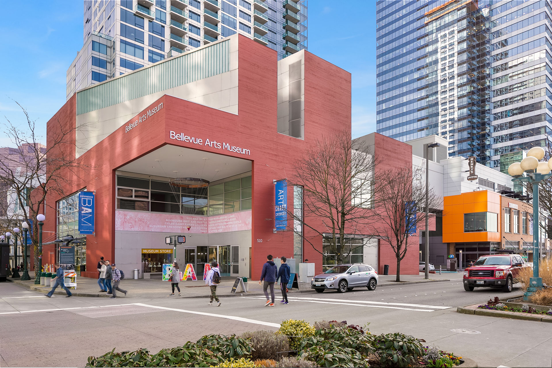 Street view of Bellevue Arts Museum, a modern red-brick building in an urban setting. People walk and cars drive by the entrance, which has a large awning. Blue banners display the museum's name. Tall buildings surround the area under a partly cloudy sky.