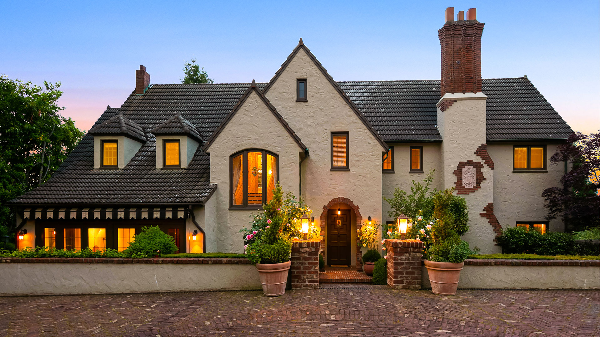 A charming two-story house with a steeply pitched roof, large chimney, and arched entrance. The exterior is adorned with various sized windows, lit warmly from the inside. The front is decorated with potted plants and illuminated lantern lights.