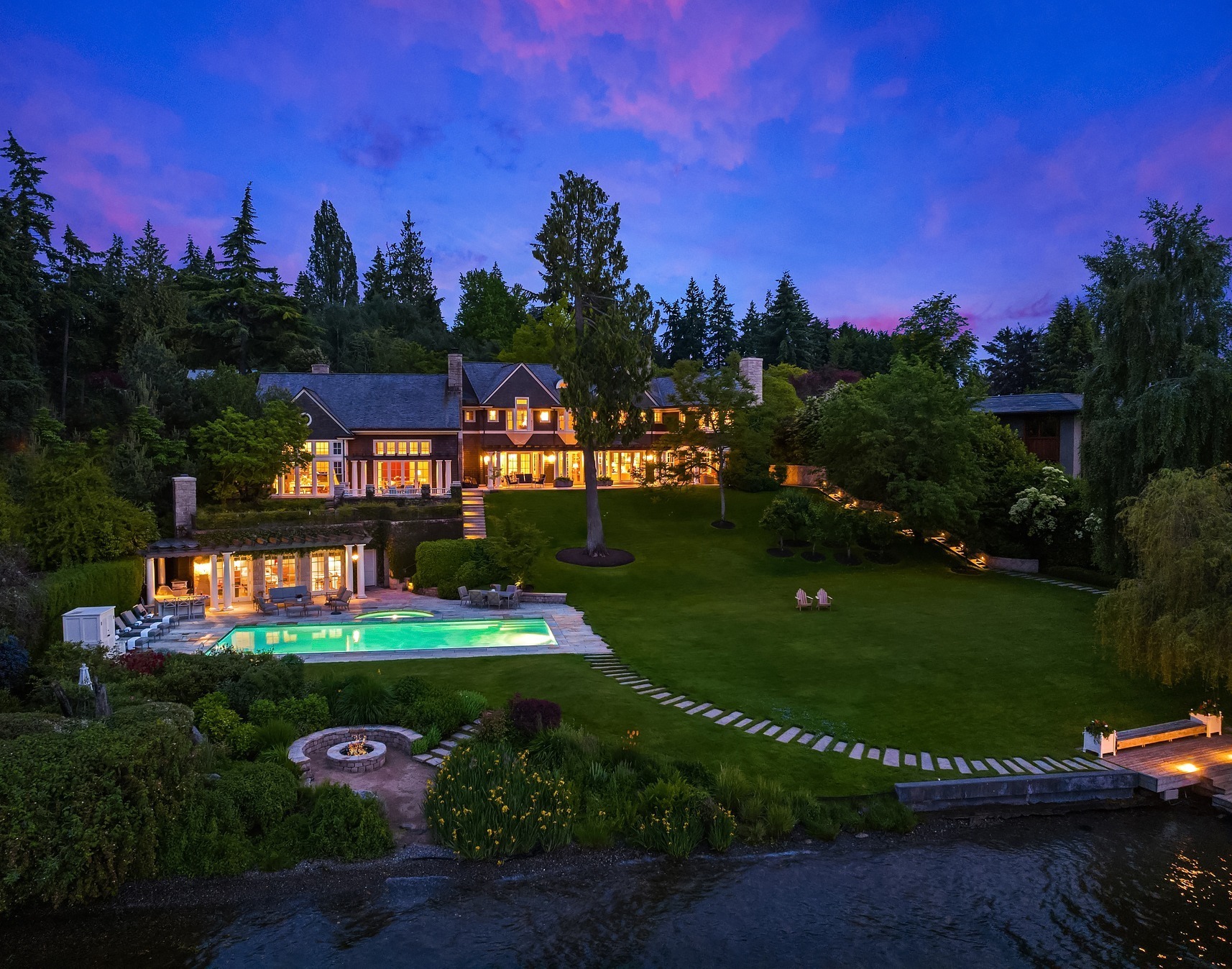 Aerial view of a luxurious lakeside mansion with expansive, well-manicured lawns, a large swimming pool, and multiple outdoor living spaces. The house is nestled among trees, and the sky is beautifully lit with colors of dusk, creating a serene and picturesque setting.