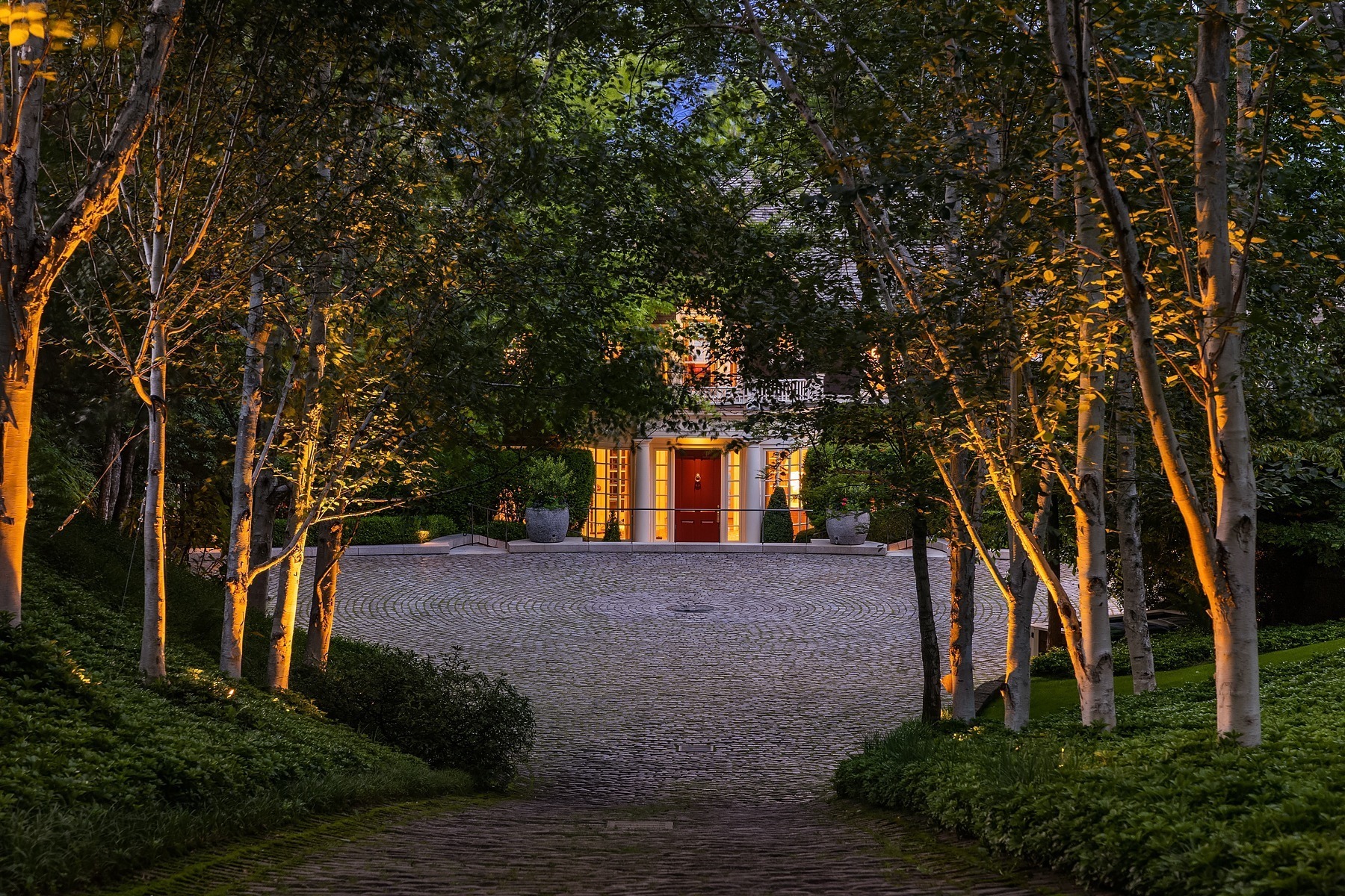 A cobblestone driveway leads through a lush, tree-lined pathway to a brightly lit house with a prominent red door. Tall trees on either side and soft ground lighting create a serene and welcoming atmosphere.