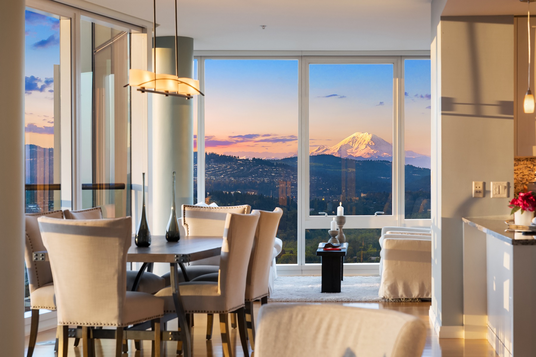 A modern dining room with a table and beige chairs is bathed in natural light from large floor-to-ceiling windows that reveal a stunning view of a snow-capped mountain at sunset. A sleek kitchen with pendant lighting is visible on the right.