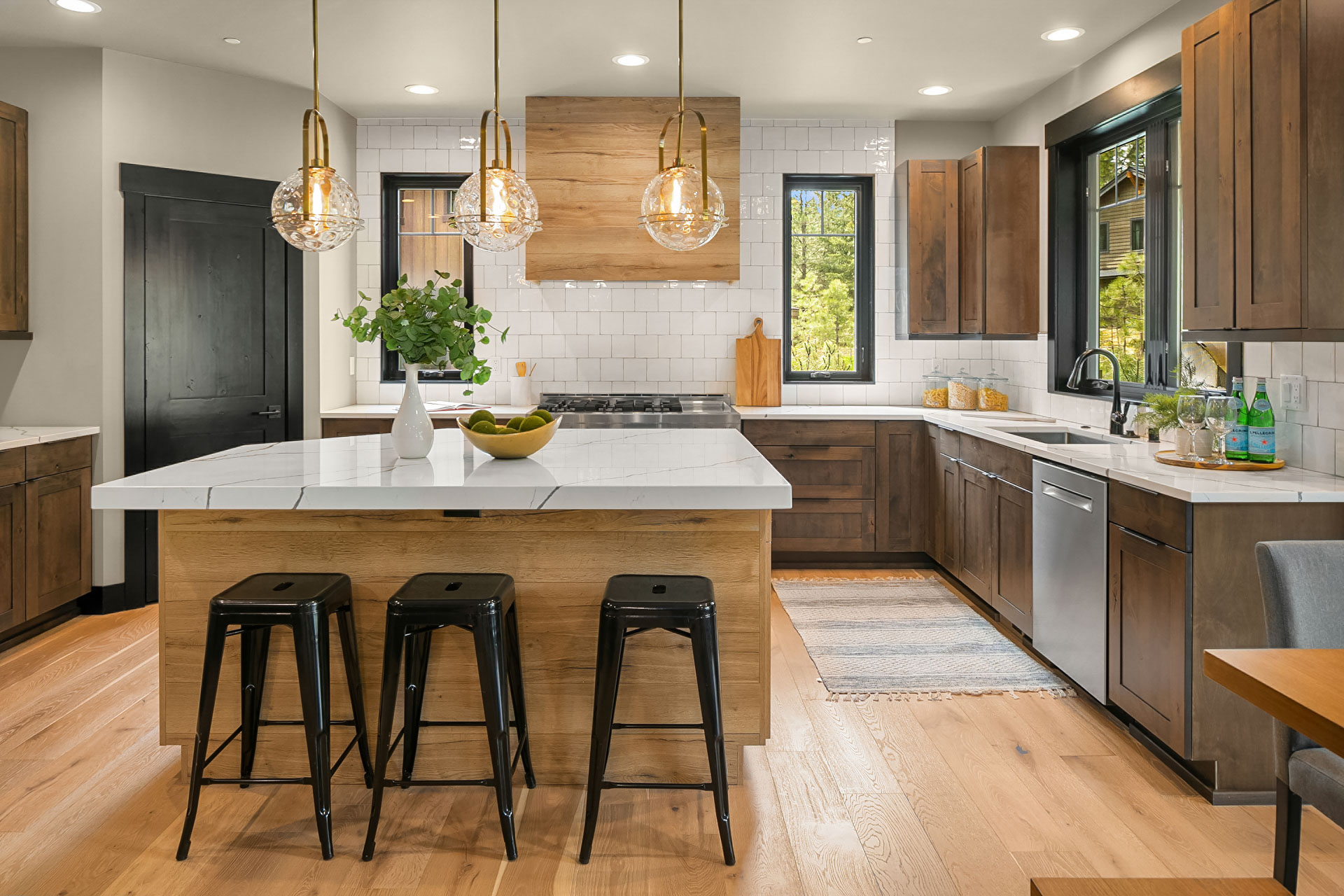 A modern kitchen features wooden cabinets, stainless steel appliances, a large island with a white countertop, and three black barstools. Three pendant lights hang above the island. A bowl of green apples and a plant decorate the island. Light-colored wood flooring accents the space.