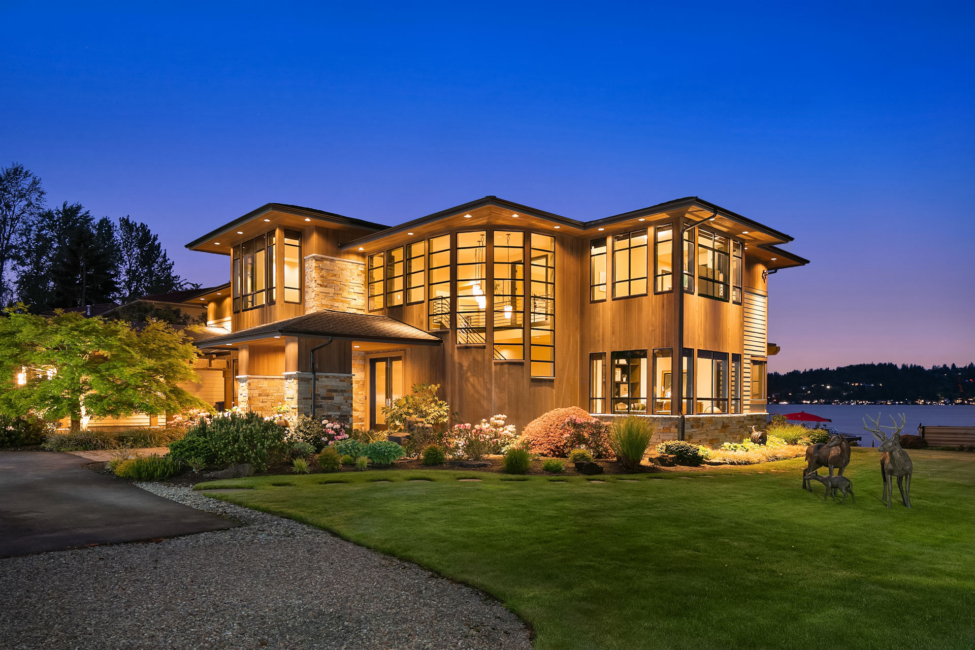 A modern, two-story house with large windows and warm exterior lighting at dusk. The property features a manicured lawn, garden with various plants and shrubs, and deer statues near the front entrance. A serene lake is visible in the background.