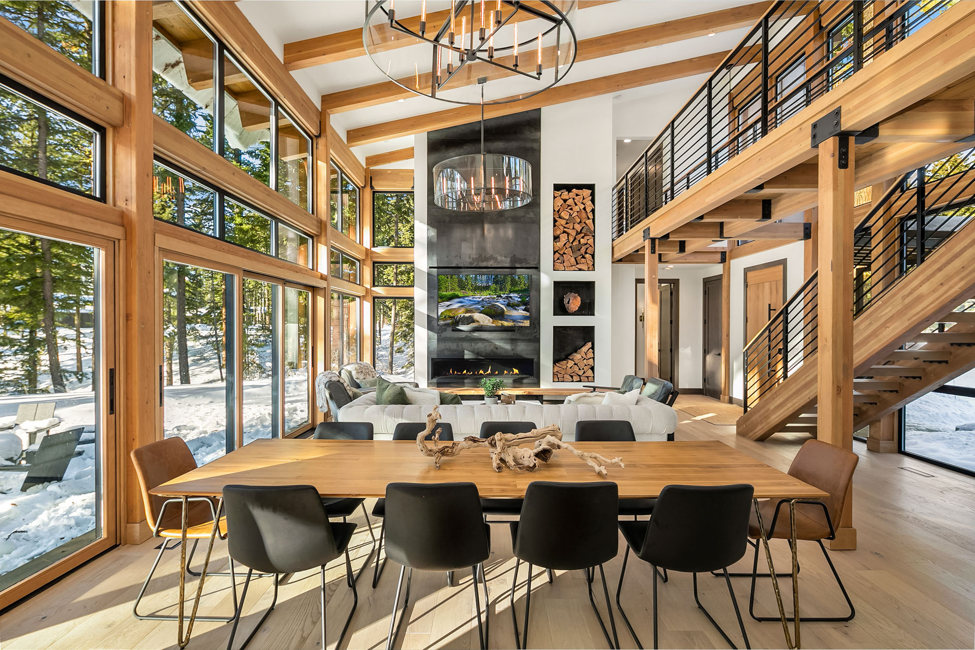 A spacious, modern living and dining area with tall windows showcasing a snowy forest. The room features wooden beams, a long dining table with black chairs, a fireplace with stacked wood, and a loft with a metal staircase. Natural light fills the space.