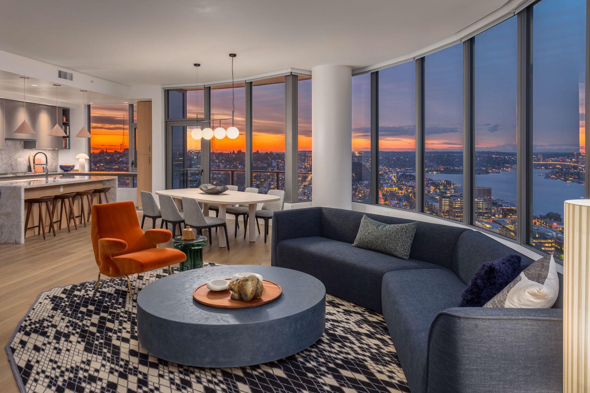 A sleek, modern open-concept living and dining area with floor-to-ceiling windows showcasing a stunning city and waterfront sunset view. The space features a comfortable gray sectional, orange armchair, round coffee table, dining set, and a contemporary kitchen.