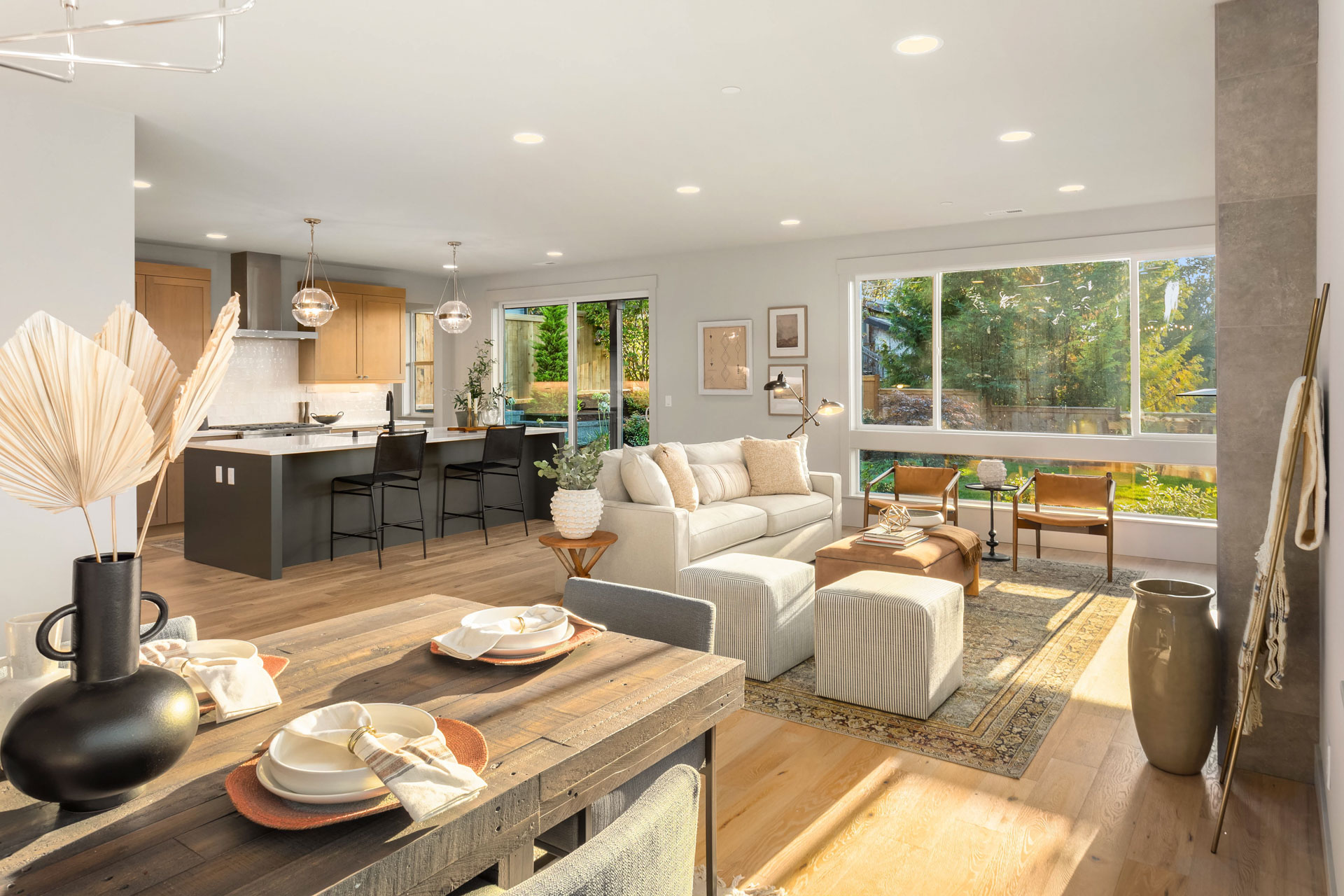 A modern open-concept living and dining area with natural light streaming through large windows. The space features a cozy sofa, a wooden dining table set for four, potted plants, and a kitchen with light wood cabinets and a breakfast bar with black stools.