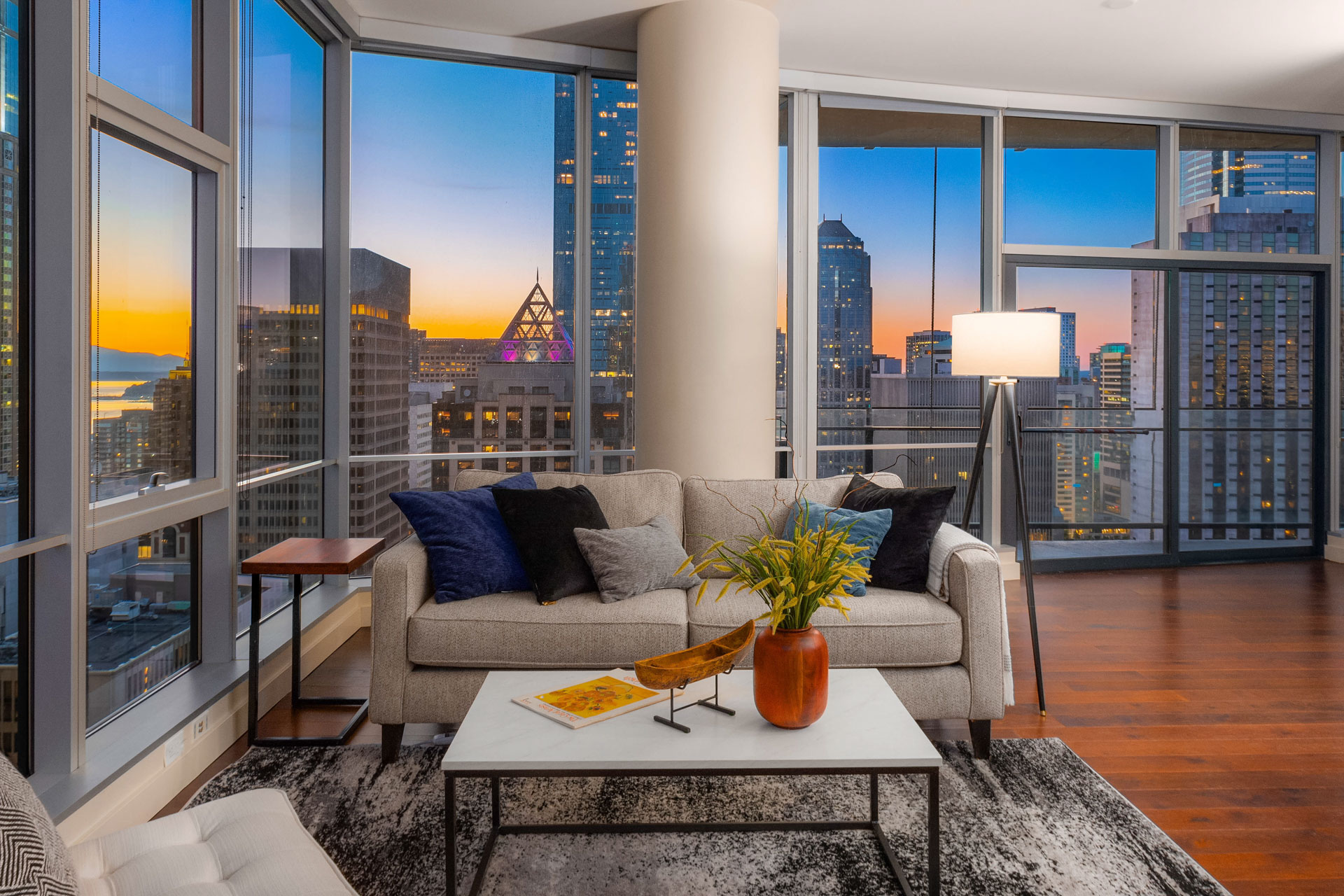 Modern living room with a large couch, decorative pillows, a coffee table, and a floor lamp. The room features floor-to-ceiling windows showcasing a cityscape at dusk with lit skyscrapers. The room combines contemporary furniture and wooden flooring.