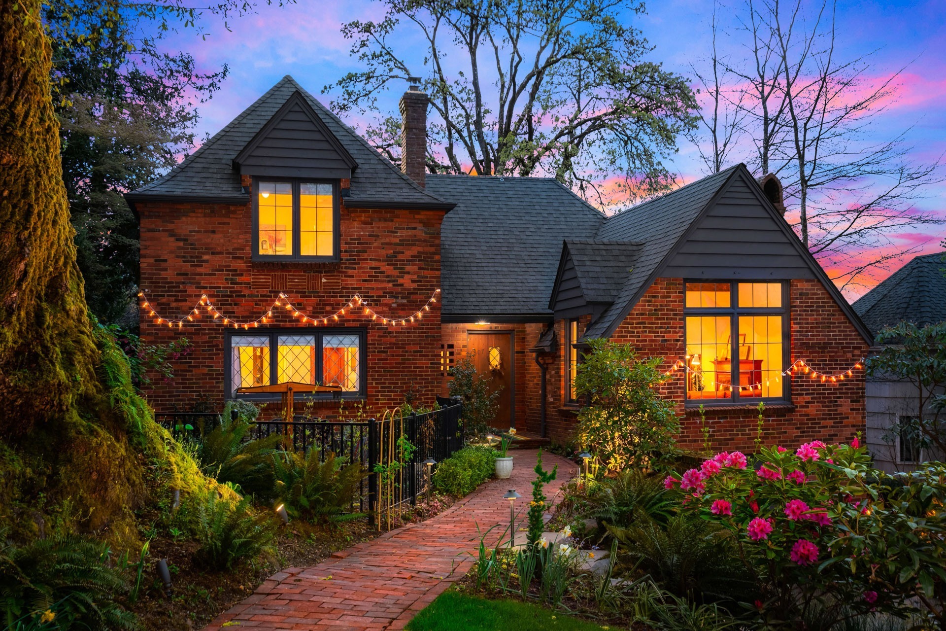 A charming brick house illuminated by warm interior lights at dusk. The exterior features white string lights, a steep roof, and decorative windows. A vibrant garden and a brick pathway lead to the front porch, surrounded by lush trees and colorful flowers.