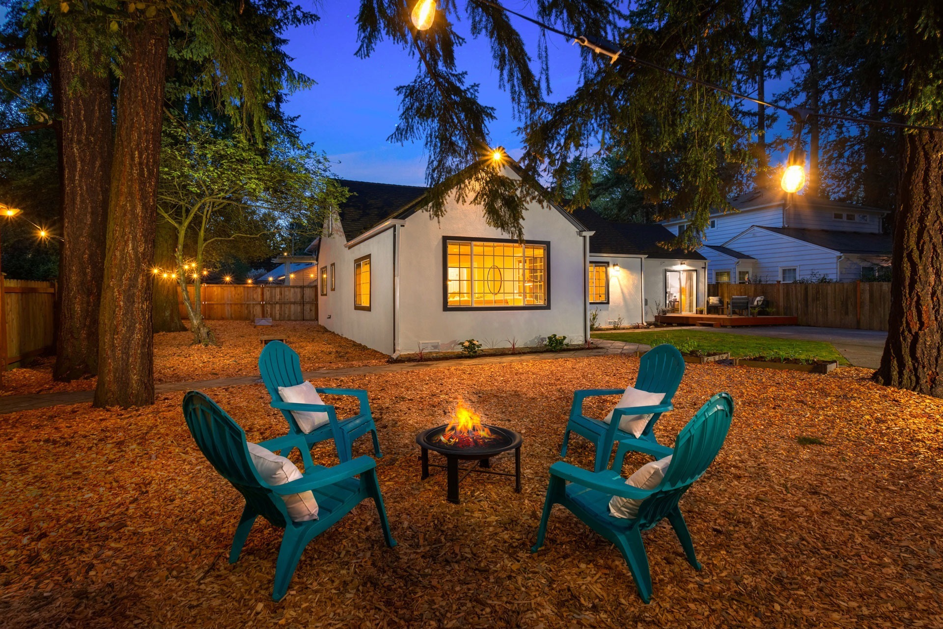 A cozy backyard scene at dusk with a small white house in the background. Four turquoise Adirondack chairs surround a small fire pit, which is lit with a small fire. Warm string lights hang between tall trees, creating a welcoming and serene atmosphere.