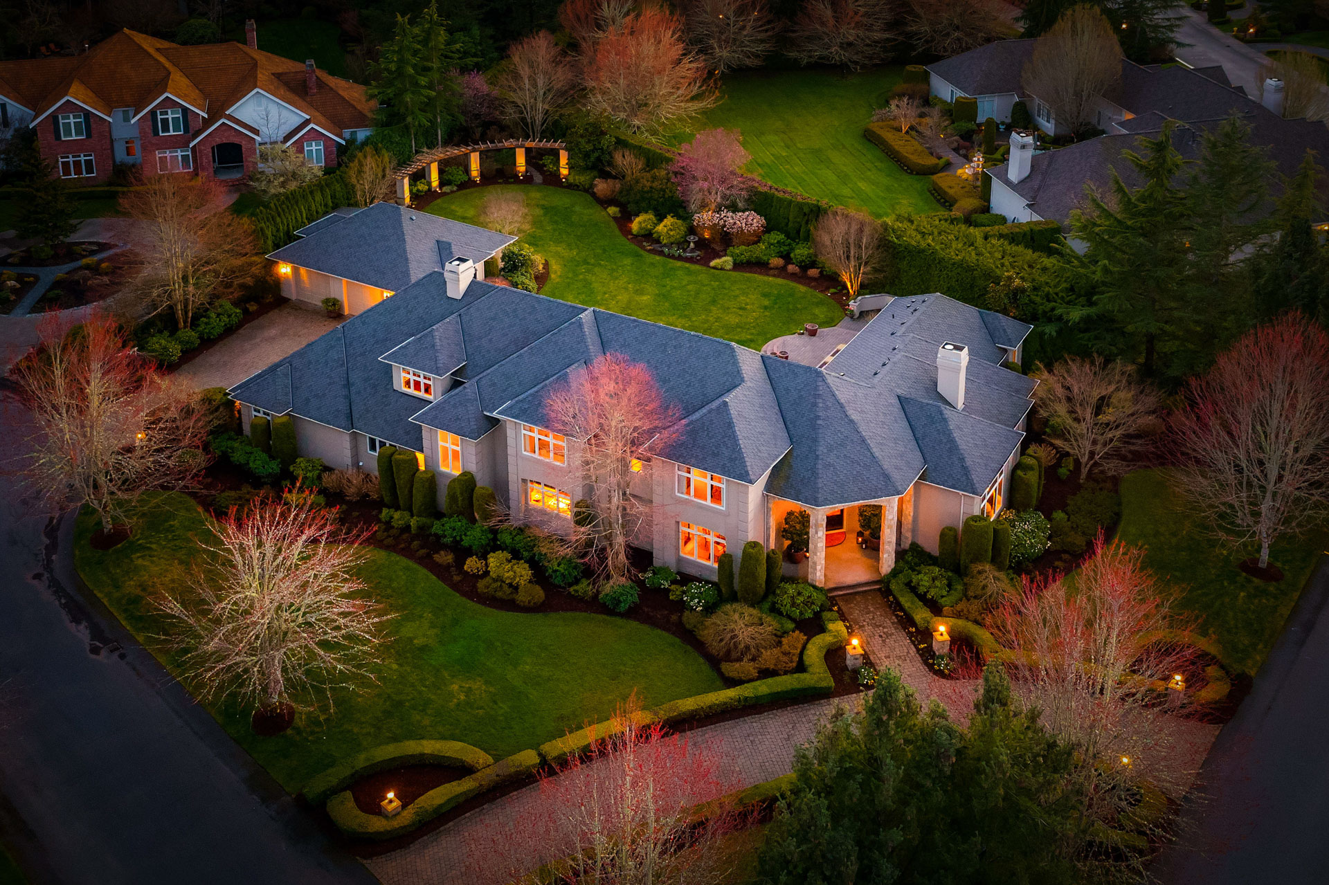 Aerial view of a large, elegant home at dusk, with warm interior lights glowing. The house is surrounded by manicured lawns, landscaped gardens, and trees with autumn foliage. Neighboring houses and a road are visible, adding to the picturesque residential scene.