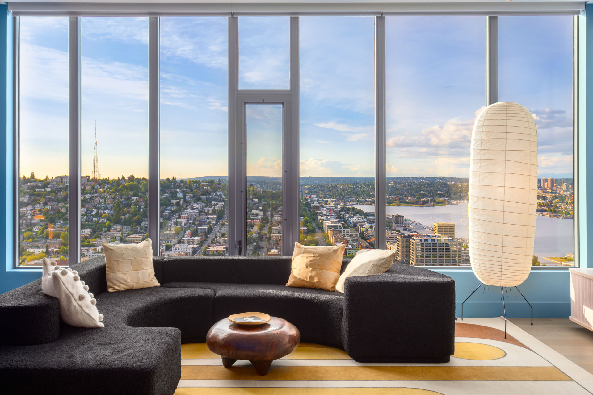 A modern living room with a black sectional sofa, beige cushions, and a wooden coffee table overlooks a cityscape through tall windows. A paper lantern-style floor lamp is on the right, and the view includes buildings, water, and a distant landscape.