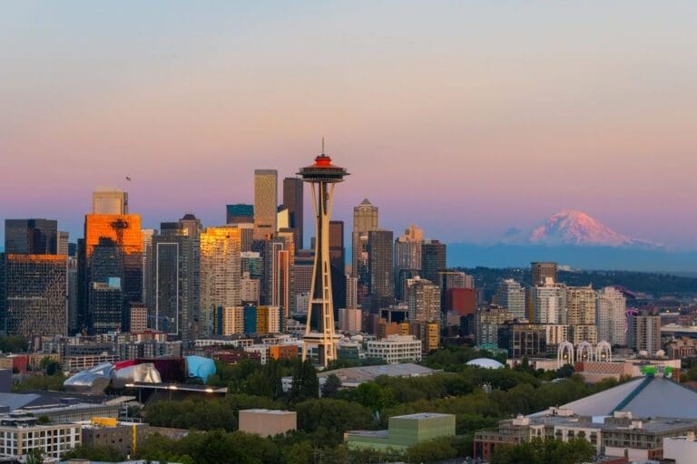 Aerial view of downtown Seattle at sunset with the Space Needle prominently in the center. Skyscrapers glow with warm light against a pastel sky, and Mount Rainier is visible in the background. Greenery and urban buildings are seen at the base.