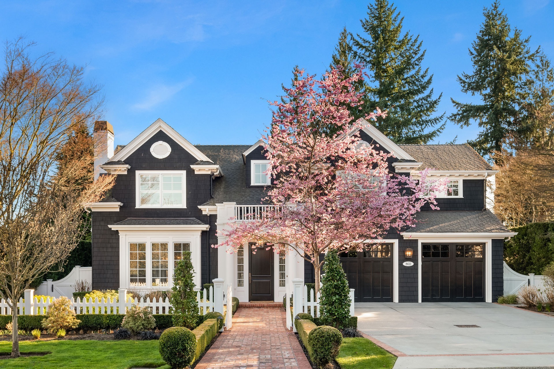 A two-story house with a dark exterior and white trim, featuring a pink blossoming tree in the front yard. The house has a brick pathway leading to the front door, symmetrical shrubs lining the path, and a white picket fence on the left side.