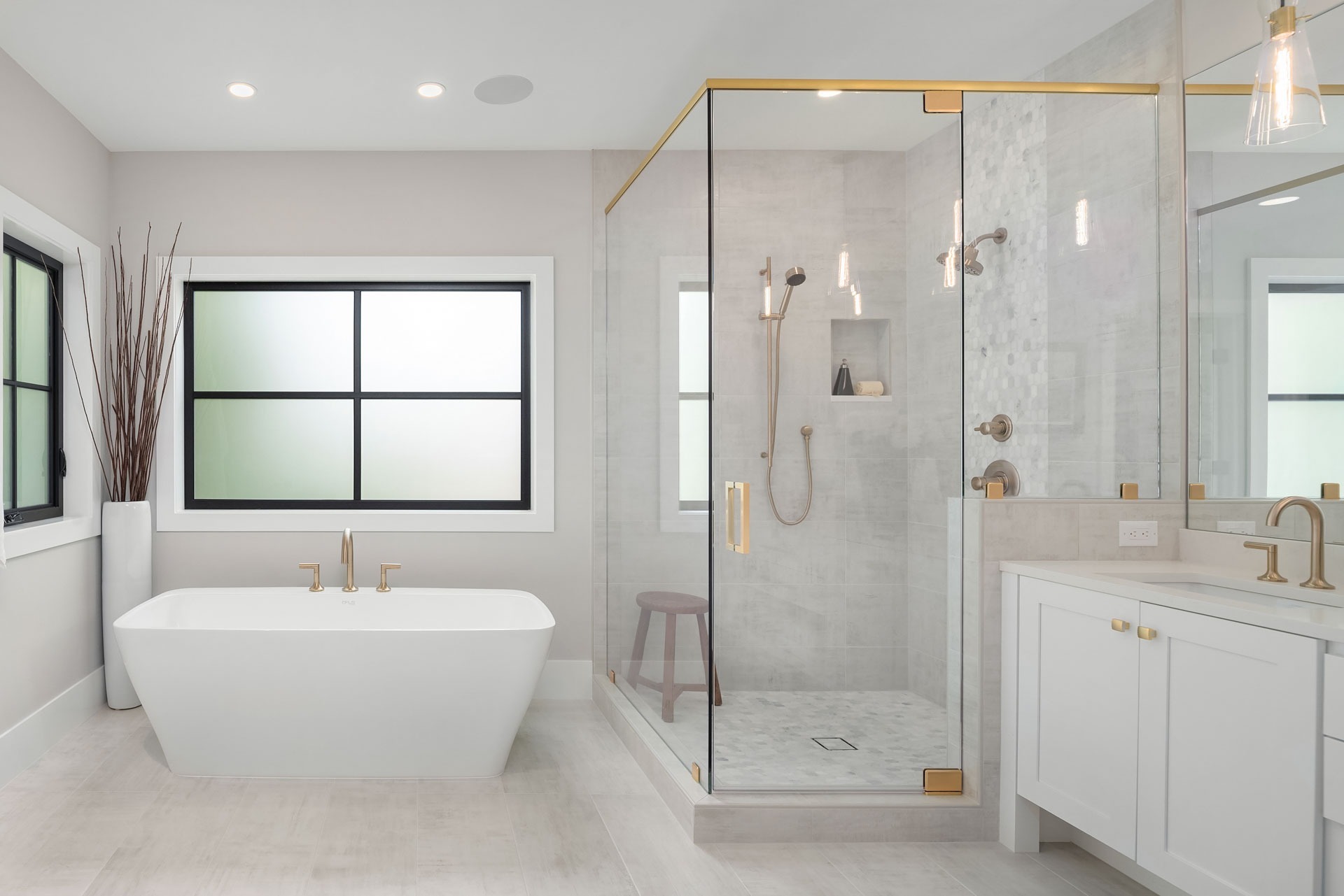 A modern bathroom featuring a glass-enclosed shower with gold fixtures, a freestanding white bathtub, and a white vanity with a gold faucet. A frosted window is behind the bathtub, and tall decorative branches are in one corner. The room has light-colored walls and flooring.