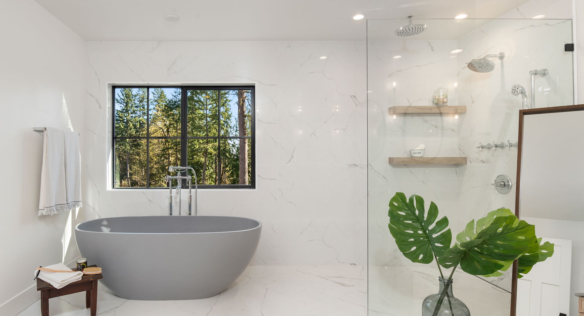 Modern bathroom with white marble walls and flooring. Room features a gray freestanding bathtub beside a large window, a walk-in glass shower with wooden shelves, tropical plant decor, a towel rack with a white towel, and a wooden side table.