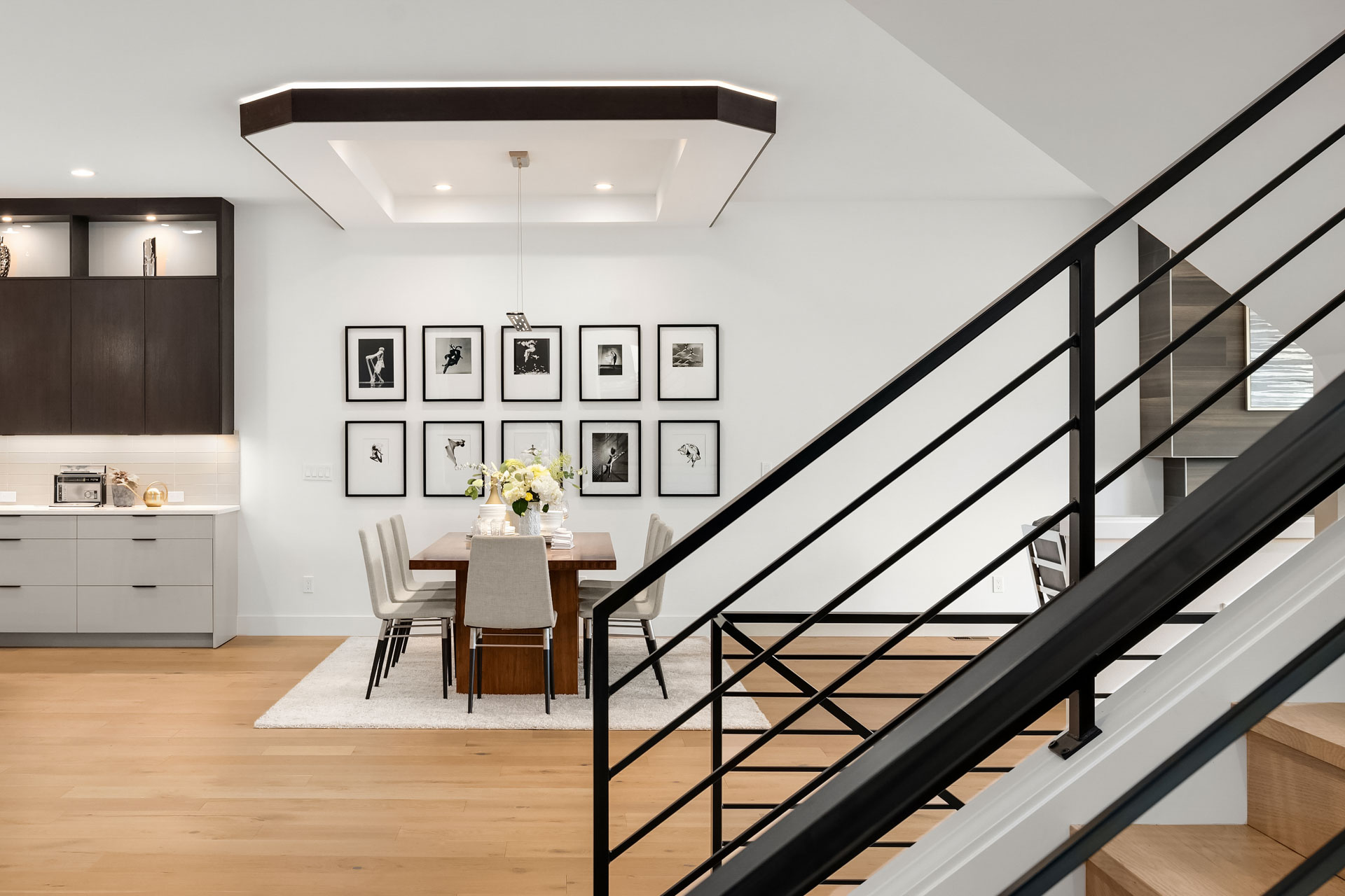 A modern dining area with a wooden table and six upholstered chairs sits in an elegant room with white walls and light wooden floors. Black and white photographs adorn the wall. A stylish light fixture hangs from a recessed ceiling, and a staircase is visible on the right.