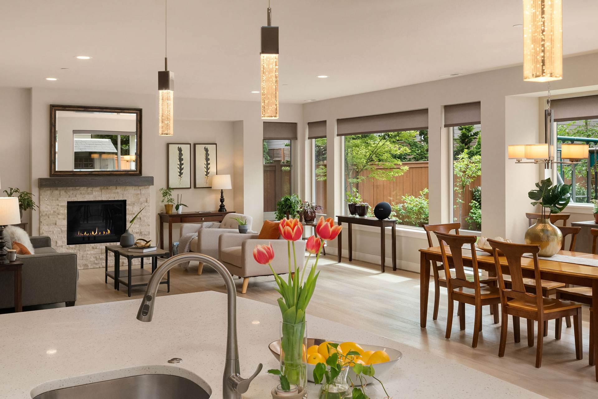 Spacious modern living room and dining area featuring light wood floors, large windows with garden views, and a stone fireplace. A kitchen island with contemporary pendant lights and a vase of tulips is in the foreground. Decor includes plants and artwork.