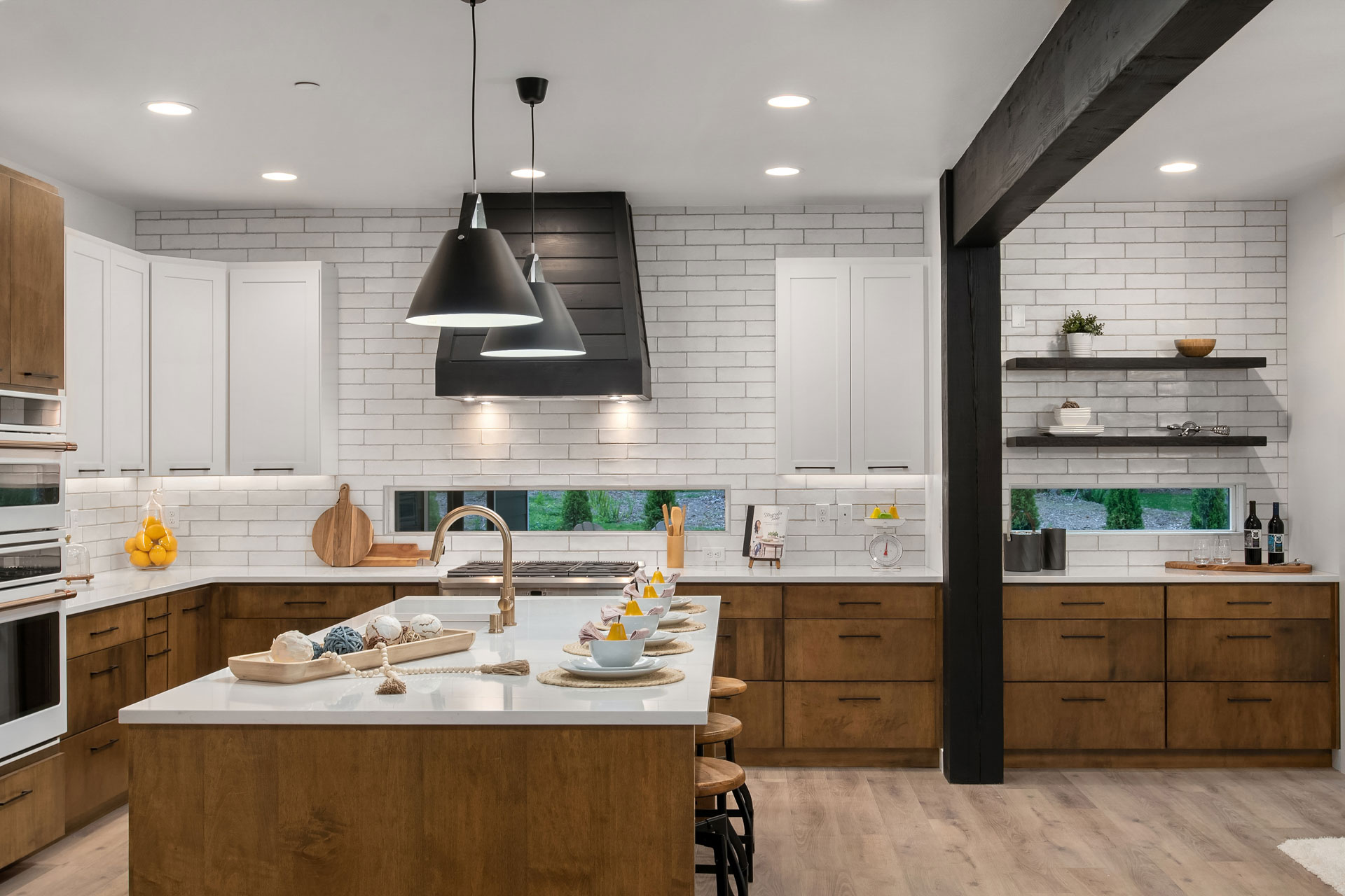 Modern kitchen with white brick backsplash, dark wood lower cabinets, and white upper cabinets. The kitchen has a large island with a sink, pendant lighting, wooden stools, and a table set with cups and bowls. Open shelves display decor items in the background.