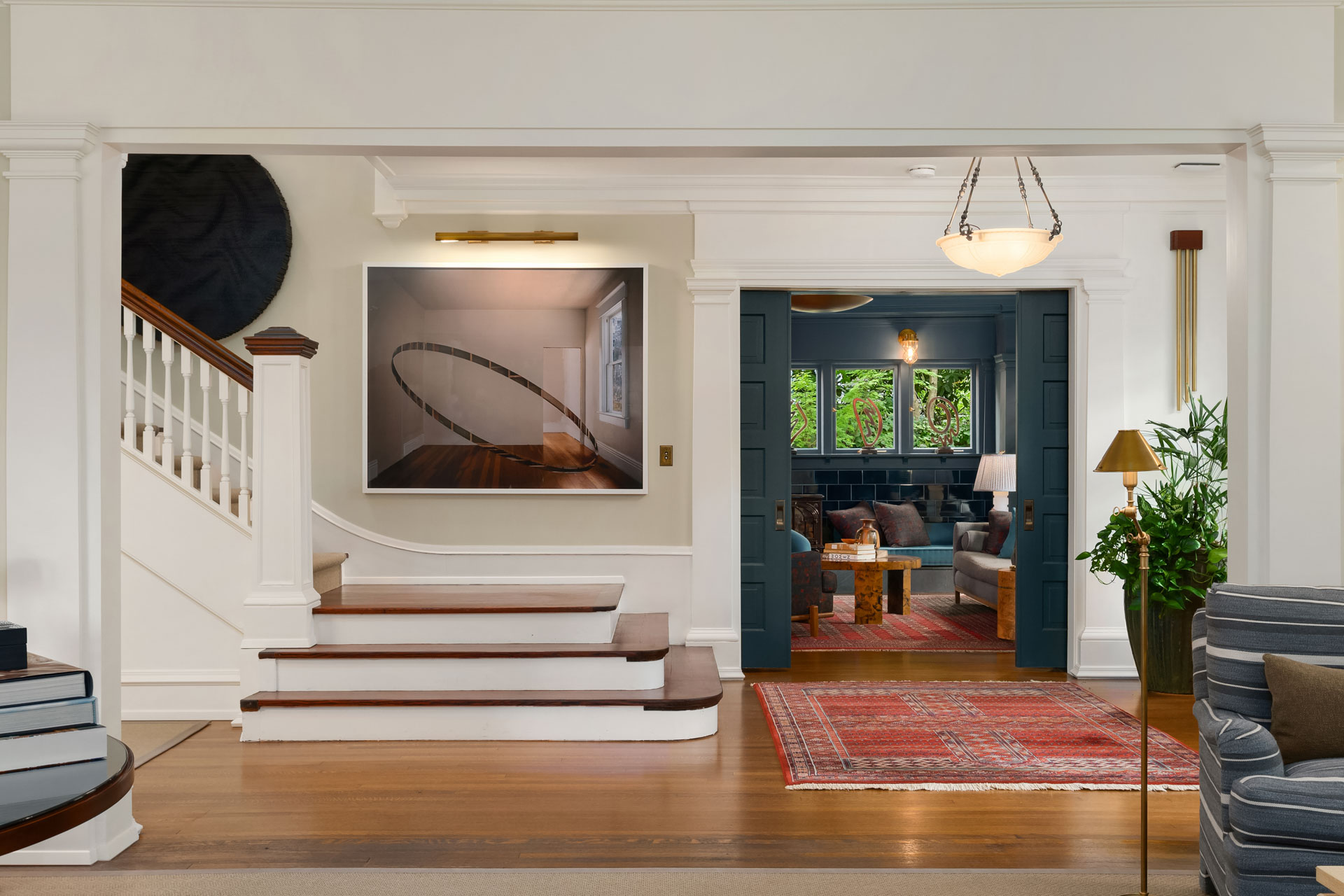 A brightly lit, open-concept living area with a white staircase and wooden steps on the left, a large piece of wall art above, and a wide doorway leading to a cozy living room. The living room features a red rug, blue walls, and a large window with greenery outside.