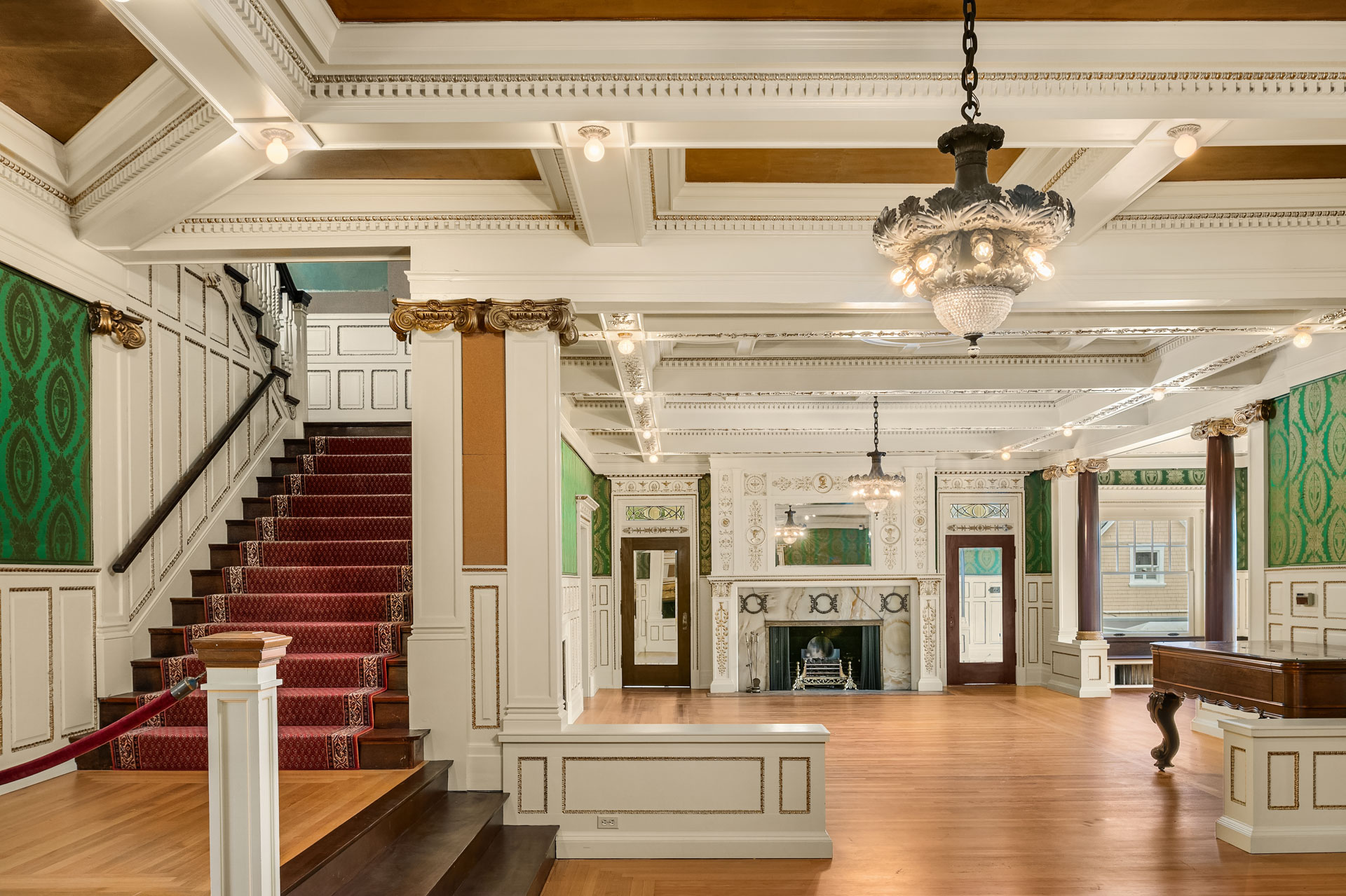 A spacious, elegant room with high ceilings and ornate moldings, featuring a grand staircase with red carpeting on the left side. The room has wooden floors, chandeliers, green patterned wallpaper, and a fireplace with a decorative mantelpiece.