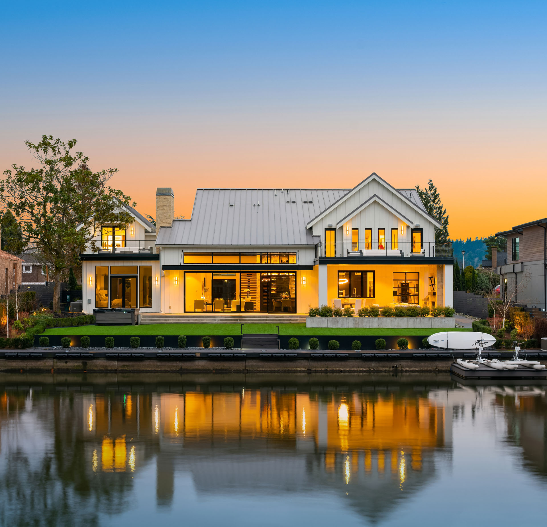 A modern white two-story house with large windows, illuminated warmly at dusk. It features a manicured lawn and direct access to a calm body of water, with a dock and a boat. The sky has a gradient of blue to orange hues as the sun sets.