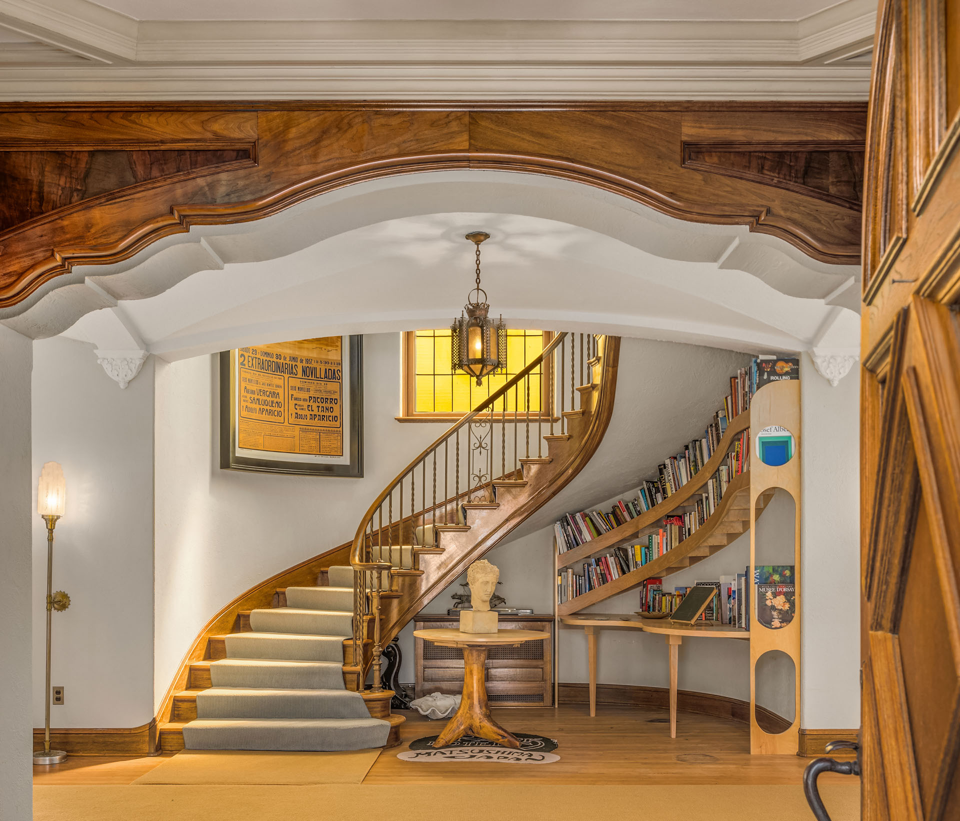 A cozy interior with a curved wooden staircase, carpeted steps, and a decorative archway. The area features bookshelves lining the wall, a round table with a bust, a vintage wall sconce, and a hanging lantern-style light. A stained glass window adds character.