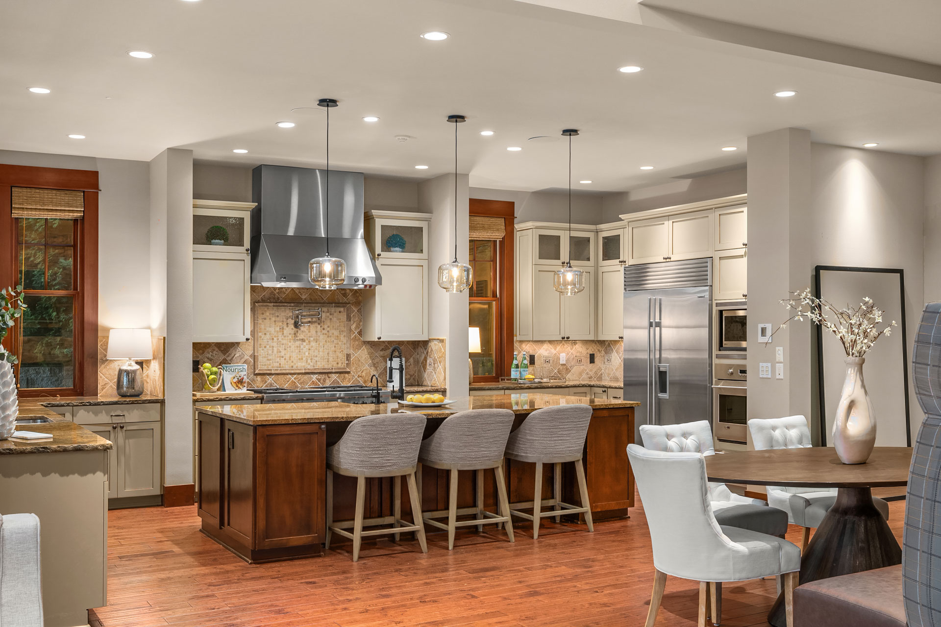 Modern kitchen and dining area featuring a central island with four cushioned bar stools, stainless steel appliances, and a tiled backsplash. Pendant lights hang above the island. The dining table is set with six chairs and decorative elements. Warm wood flooring throughout.