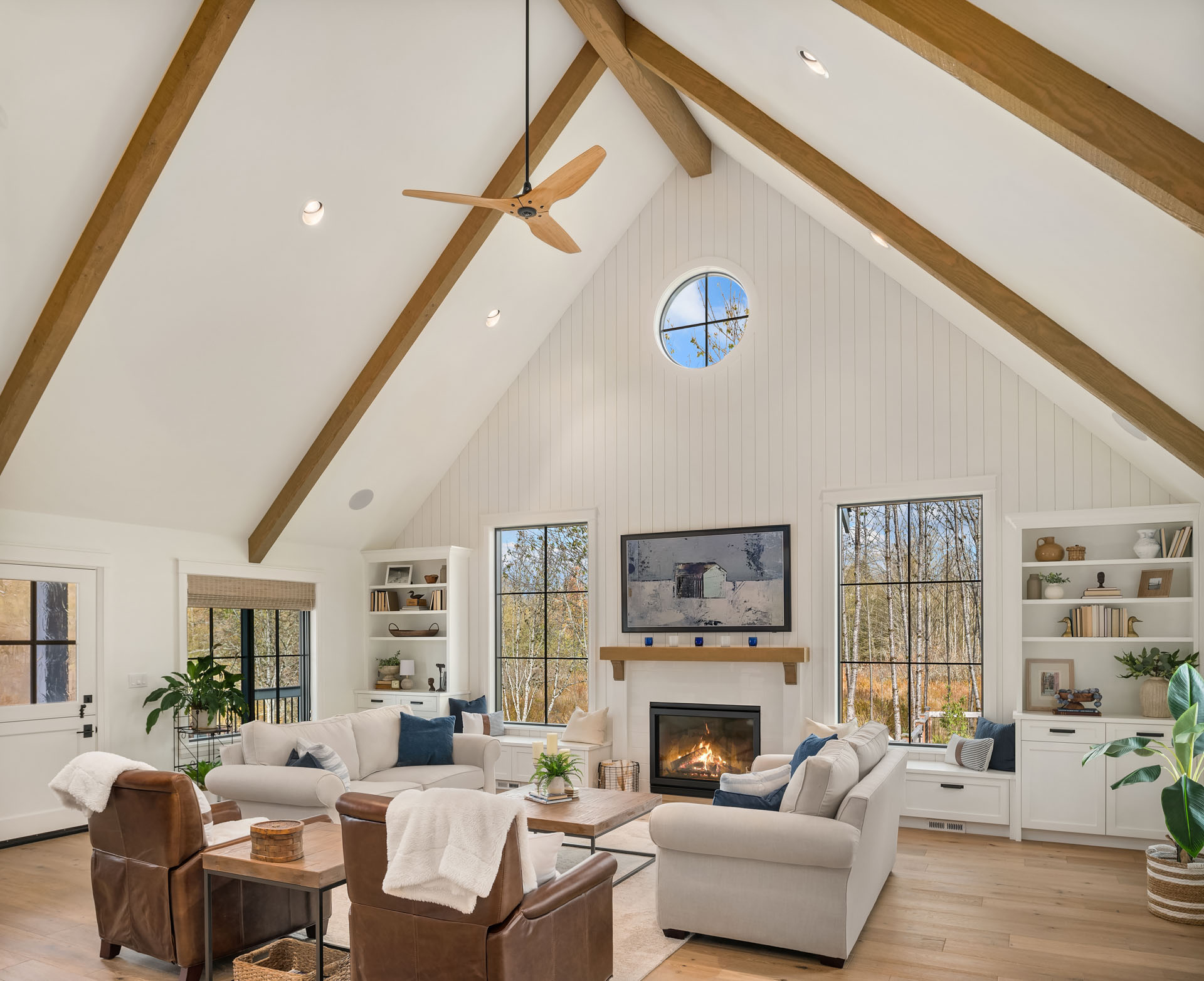 A spacious living room with high, vaulted ceilings featuring wooden beams. The room has a cozy seating arrangement with white sofas around a lit fireplace. Large windows allow natural light to fill the space, and built-in shelves hold books and decorations.